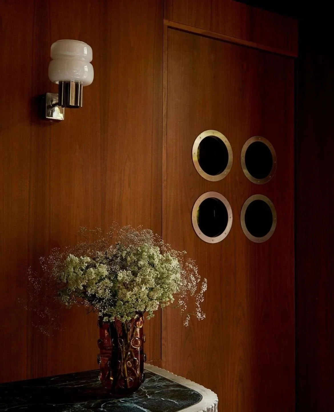 A wooden wall with four portholes, a modern wall sconce with an opaque white glass shade, and a vase with white flowers on a dark marble surface.