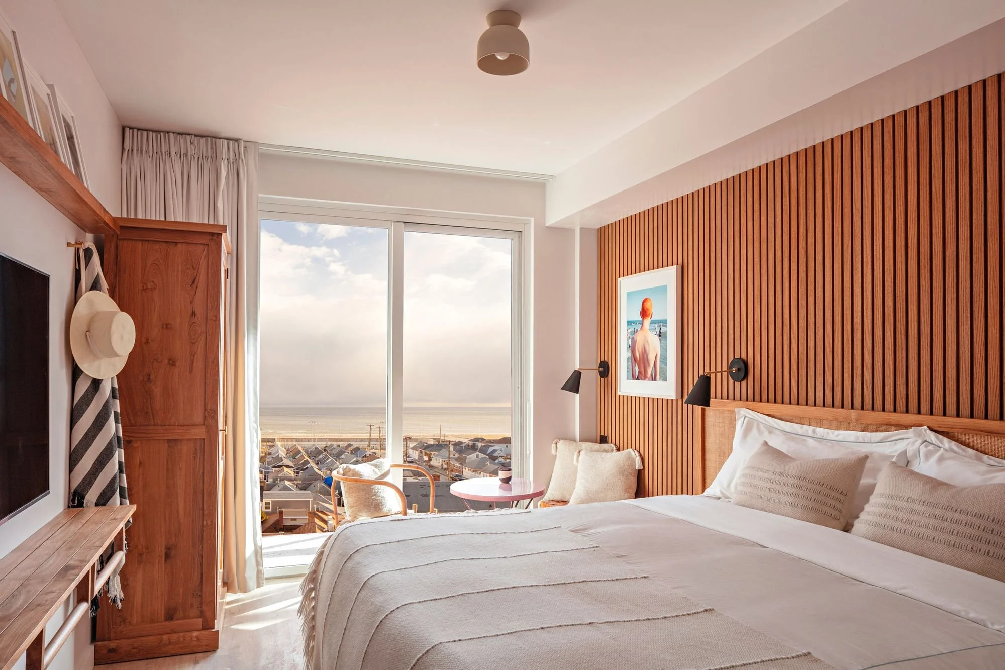 Hotel bedroom with a large bed, wooden headboard, art on the wall, black wall lamps, a window with a view of beach houses and the ocean, and a small pink table with a chair.