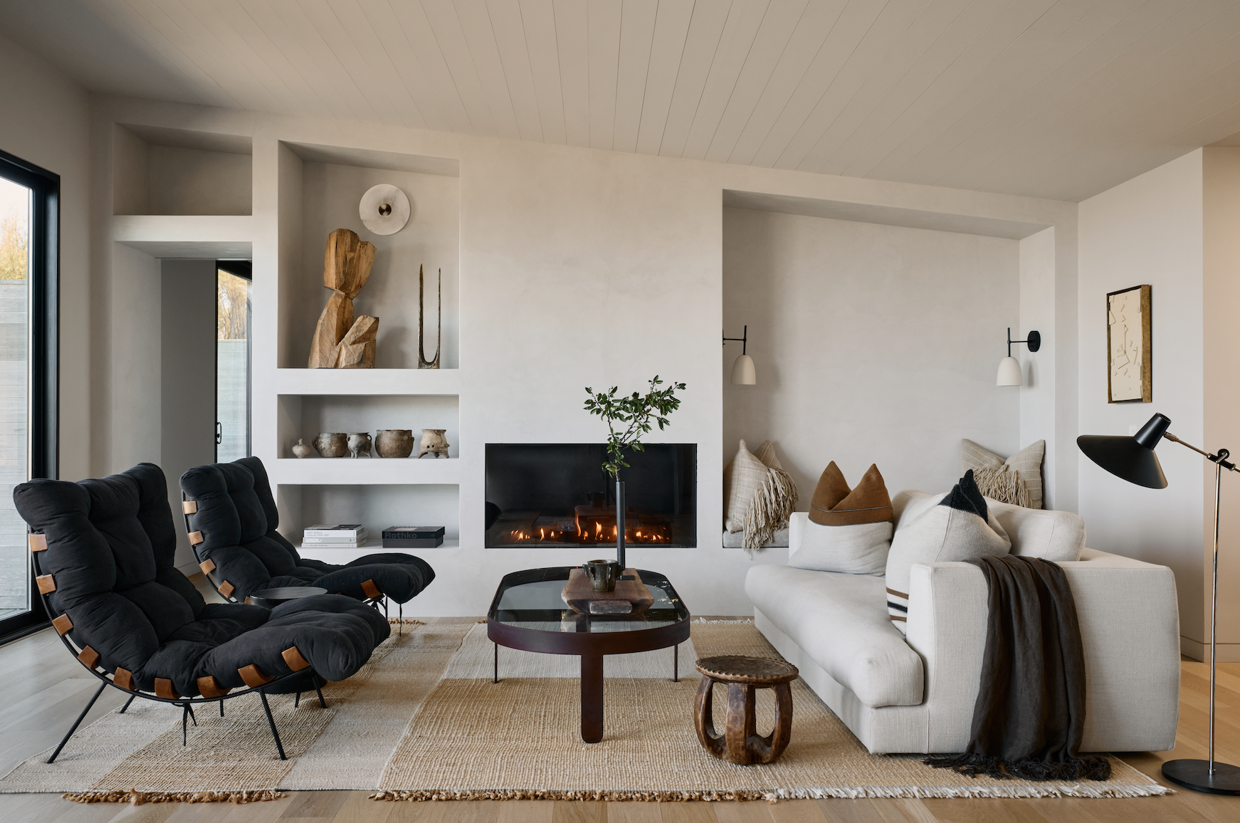 Modern living room with white walls, black lounge chairs, a white sofa, a central coffee table, and a fireplace with a black framed opening. Shelves with decorative objects are built into the wall, and there are accent pillows and a throw blanket on the sofa. A floor lamp is positioned beside the sofa.