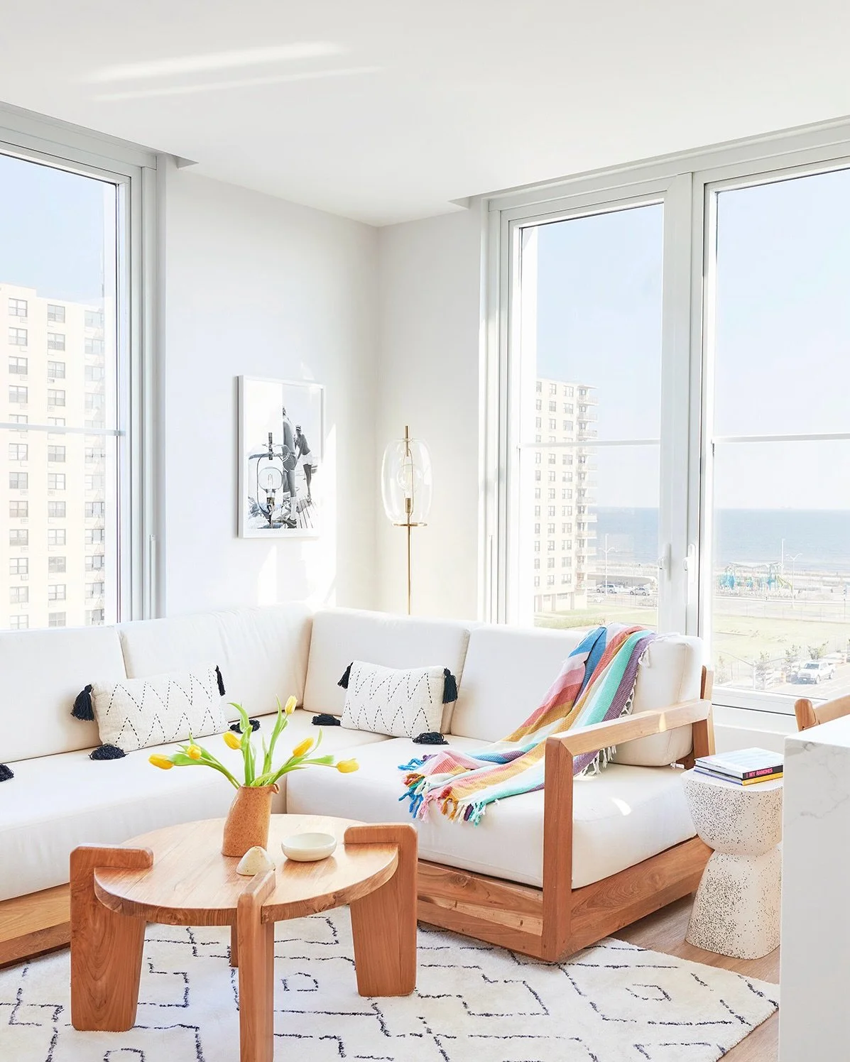 Bright living room with large windows, a white sectional sofa with decorative pillows, a colorful striped blanket, a wooden coffee table with a vase of yellow tulips, a framed black and white art piece, a standing lamp, and a view of high-rise buildings and the ocean outside.