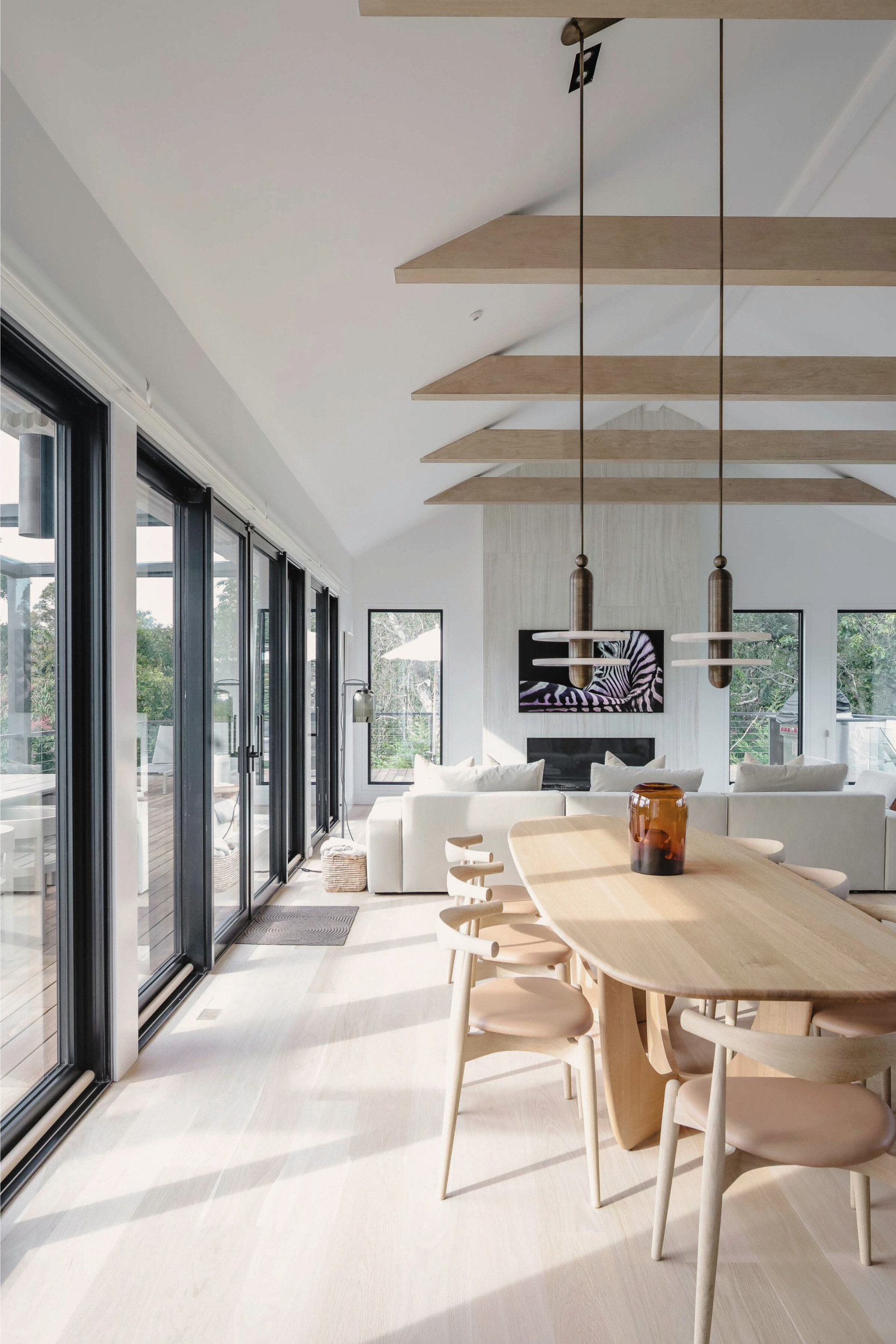 Hamptons Interior Design Bright, modern living and dining area with floor-to-ceiling sliding glass doors, white sofas, light wood dining table and chairs, a wall-mounted TV, and wooden ceiling beams.