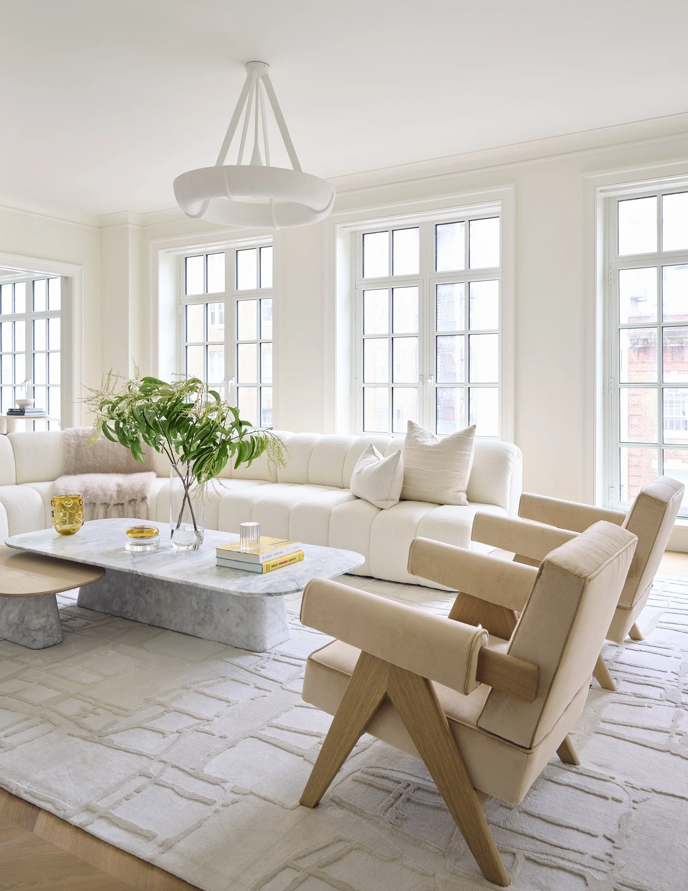 New York City Interior Design. Bright living room with white walls, large windows, beige armchairs, a white sectional sofa, a marble coffee table, and decorative items.