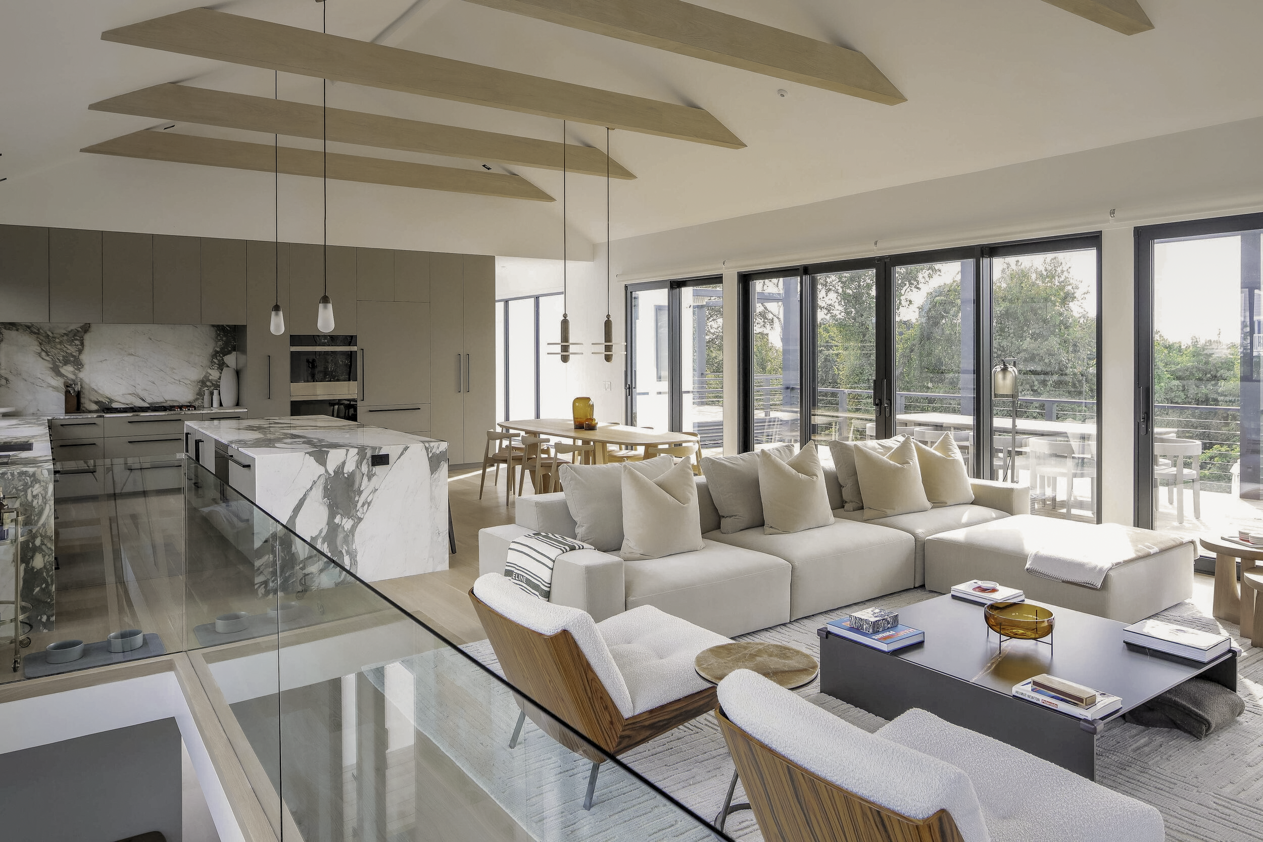 Montauk Interior Design. Open-concept living room with a white sectional sofa, wooden armchairs, black coffee table, and large glass sliding doors leading to an outdoor balcony with trees visible outside.