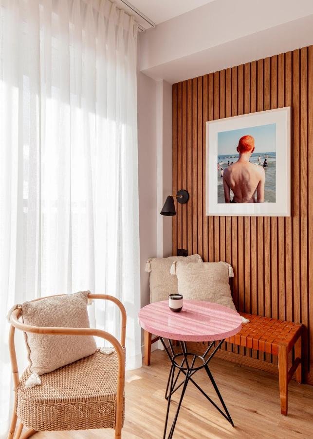 Corner of a cozy room featuring a wicker armchair with a beige cushion, a small pink round table with a black metal base holding a black and white cup, a wooden bench with cushion, a wooden-paneled wall with a framed photograph of a person with red hair at the beach, and sheer white curtains covering a large window.