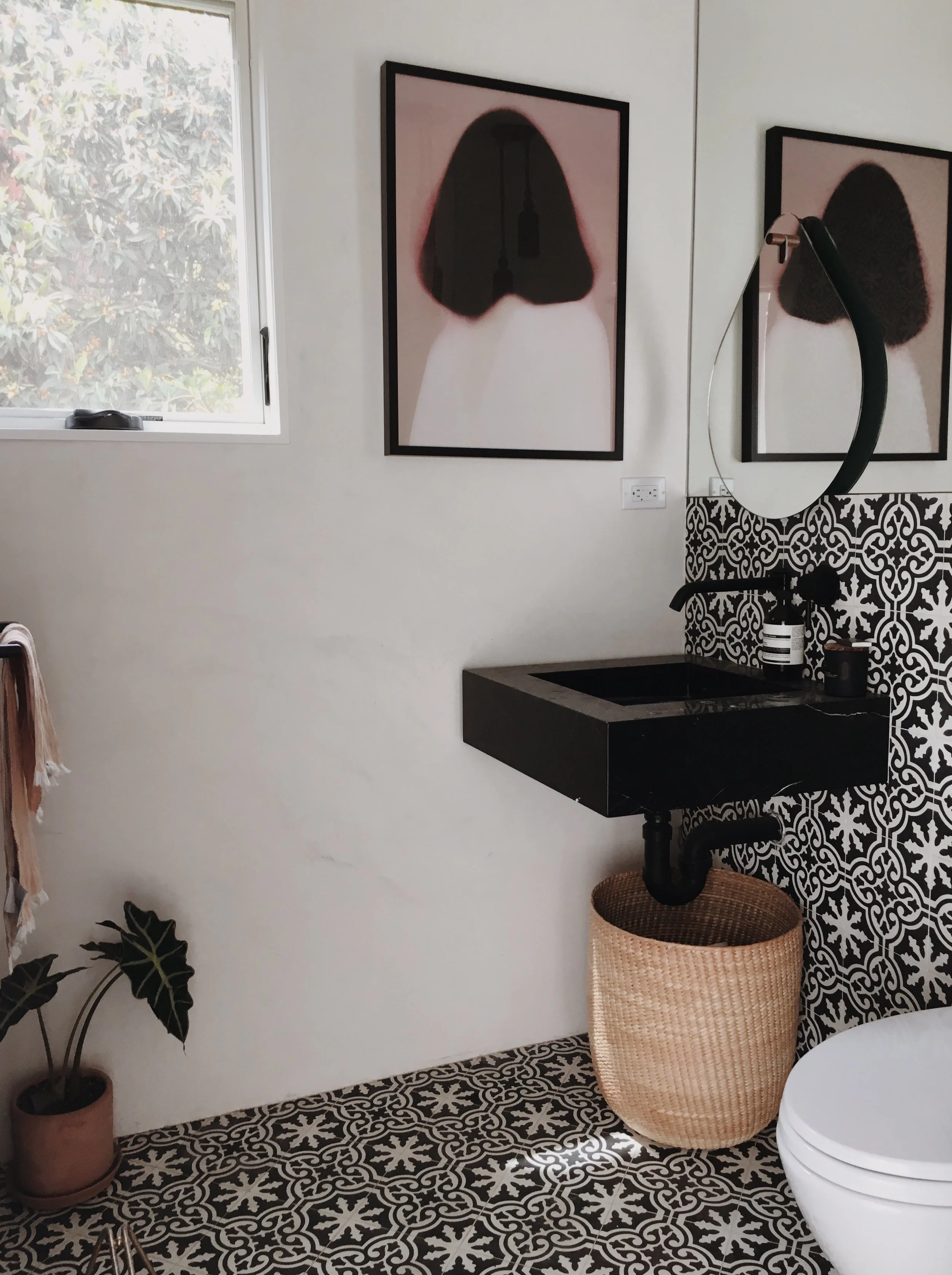 Los Angeles interior Design. Powder Room design. A modern bathroom with black and white patterned floor and wall tiles, a black sink, a circular mirror, wall art, a potted plant, and a window.