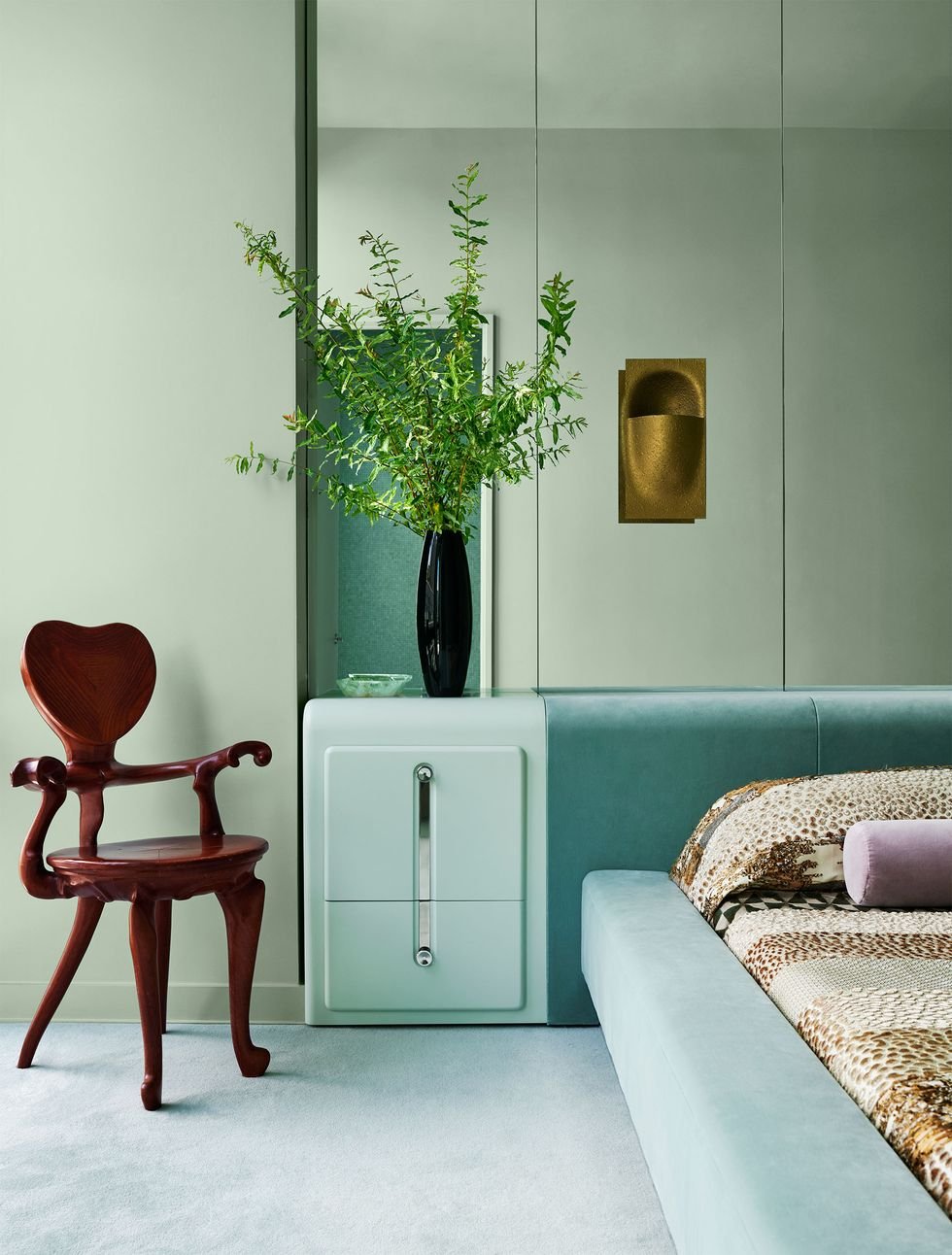 New York Interior Design. Interior of a modern bedroom with a dark wooden chair, green upholstered bed, and a large vase with green leafy branches on a white side table against a gray wall with minimalist decor.