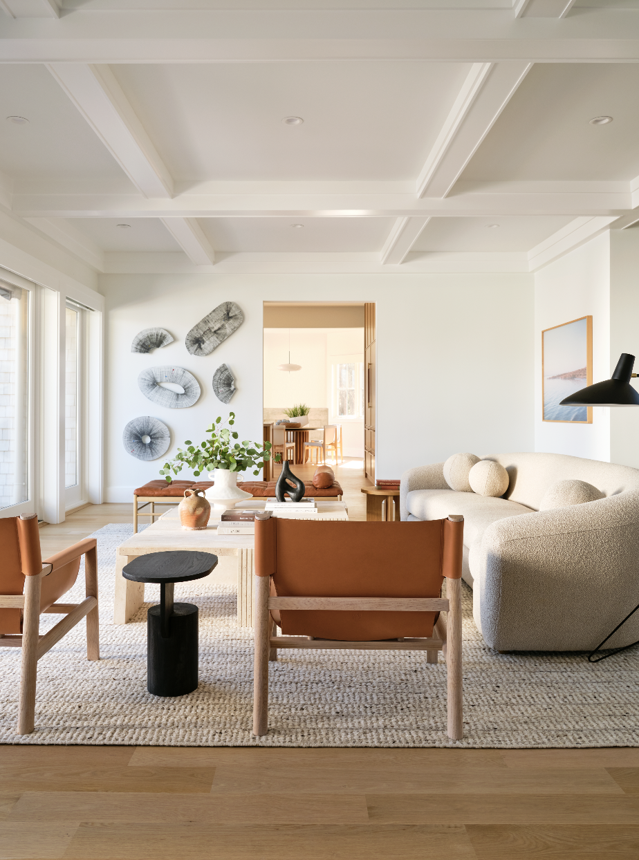 Bright living room with white walls, vaulted ceiling with beams, large windows, and light wood flooring. Contains beige sofa, wooden chairs with leather straps, a coffee table with vases and books, a side table, wall art, and wall decor. A doorway leads to a dining area with a wooden table and chairs.