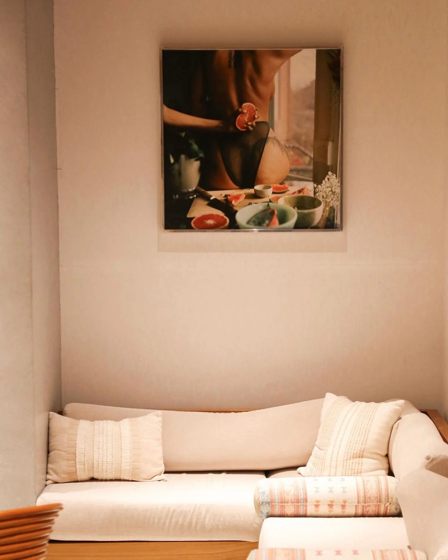 A wall with a framed artwork showing a person in black underwear preparing food, with bowls and ingredients on a table. Below the artwork is a beige couch with white and patterned cushions.
