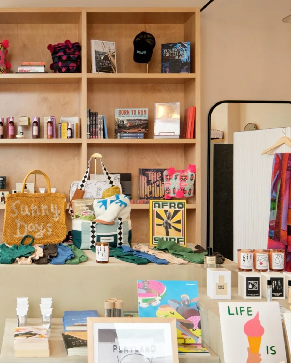 Display of colorful bags, books, artwork, and skincare products on a table and shelves in a retail store.