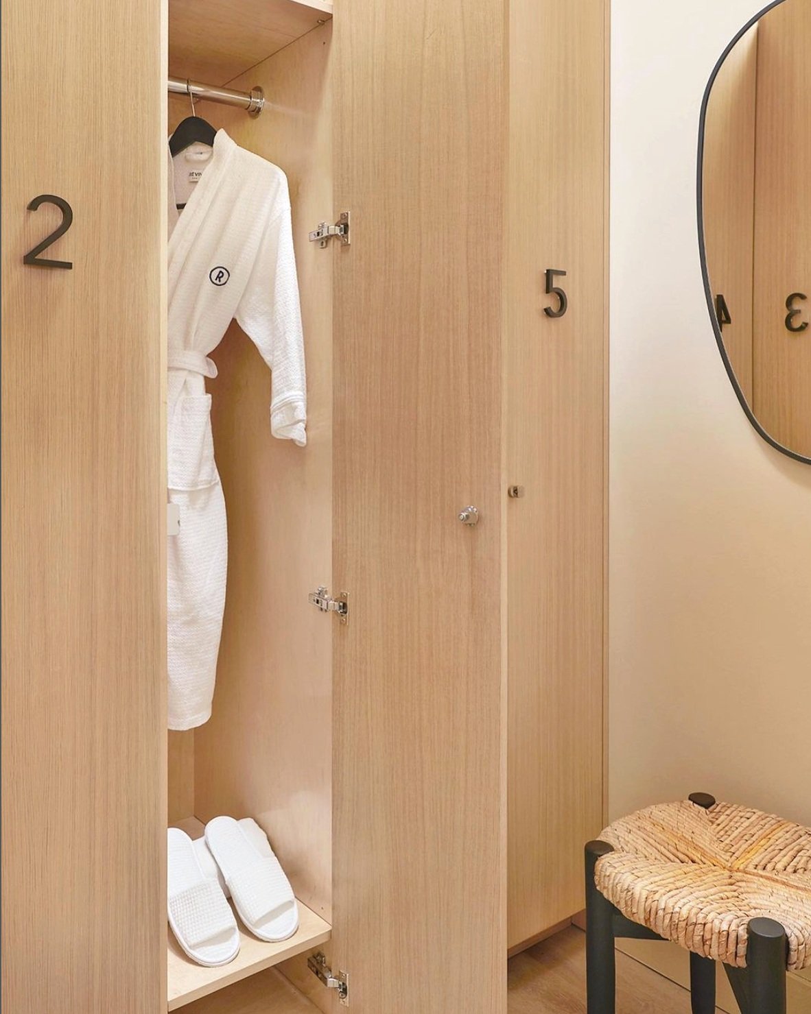 Open wooden locker with a white bathrobe and slippers inside, and a round mirror on the wall nearby.