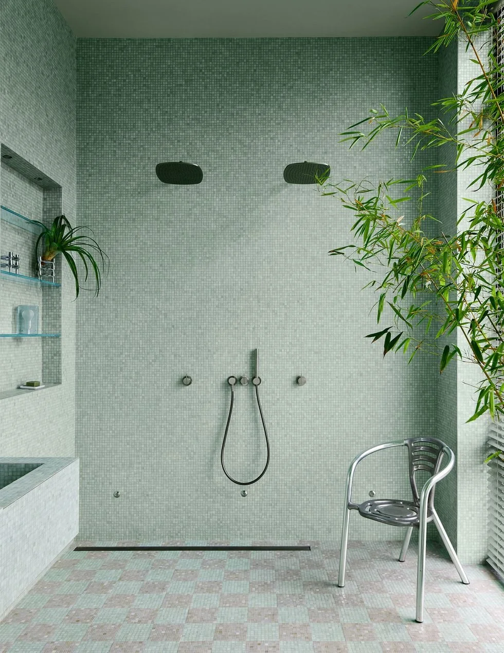 A minimalist spa shower area with light green mosaic tiles, two showerheads, a black metallic chair, and green plants.