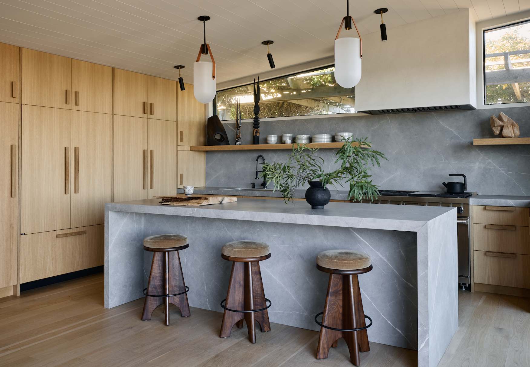 Hamptons Interior Design. Modern kitchen with wooden cabinets, a marble island with three wooden stools, a plant in a black vase, hanging pendant lights, and open shelves with dishes and decorative items.