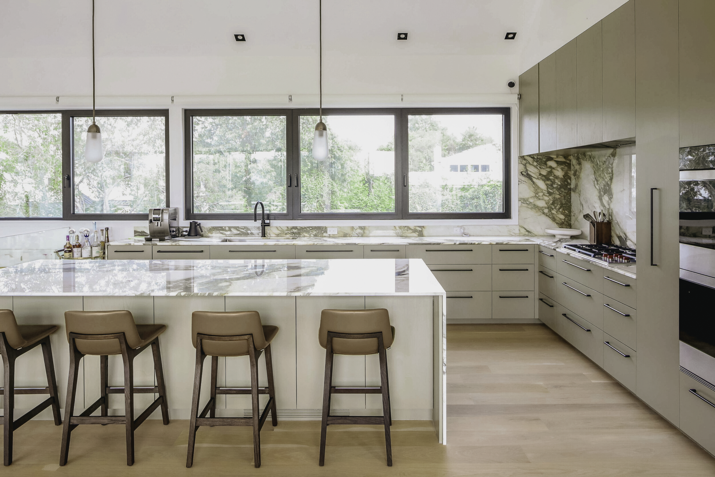 Montauk Interior Design. Modern kitchen with beige cabinets, marble backsplash, large windows, island with four brown chairs, and hardwood floors.