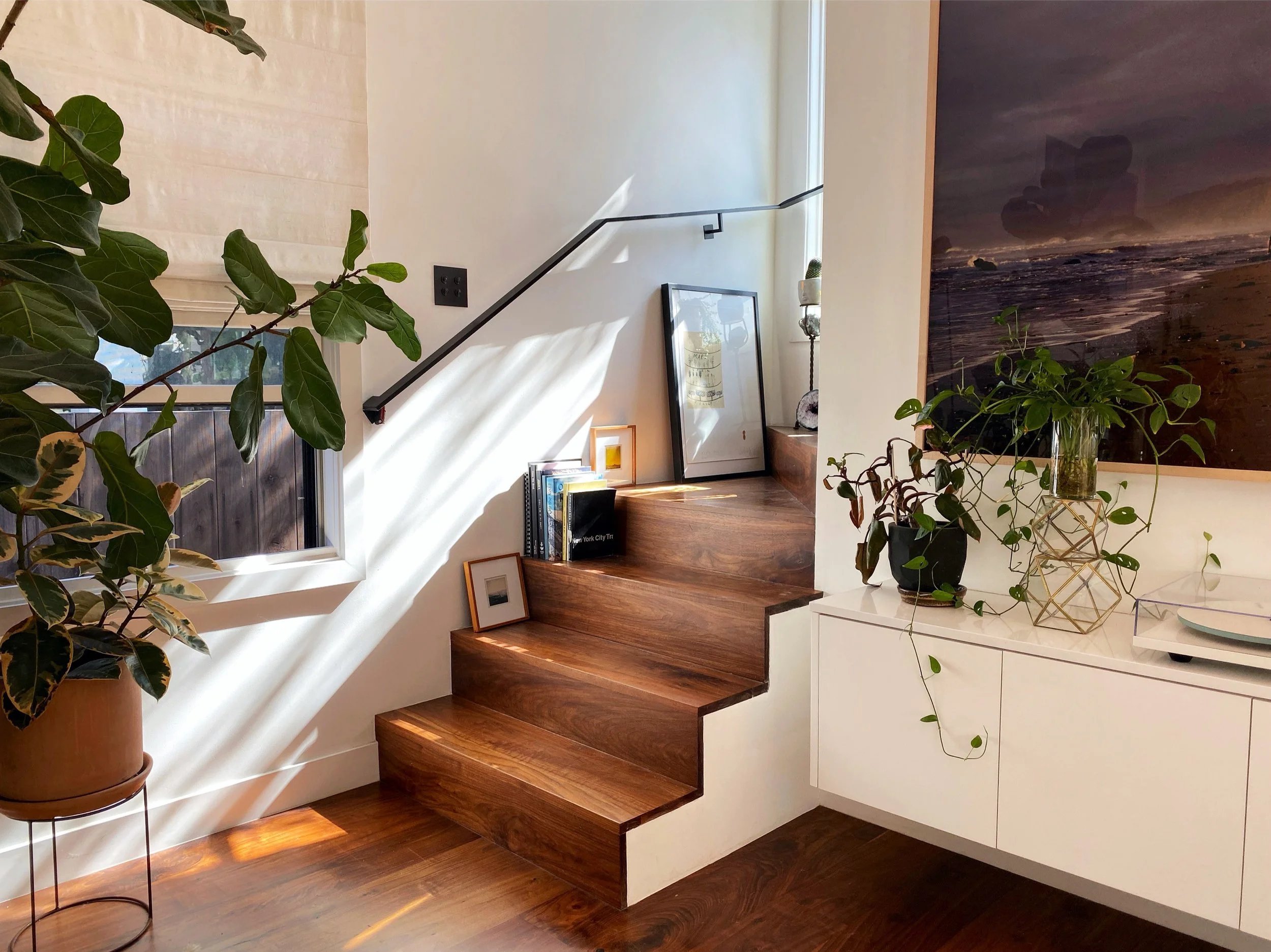 Los Angeles interior Design. Interior view of a staircase with wooden steps, surrounded by potted plants, a white cabinet with decorative items, and framed pictures. Sunlight creates shadows on the white wall and window.