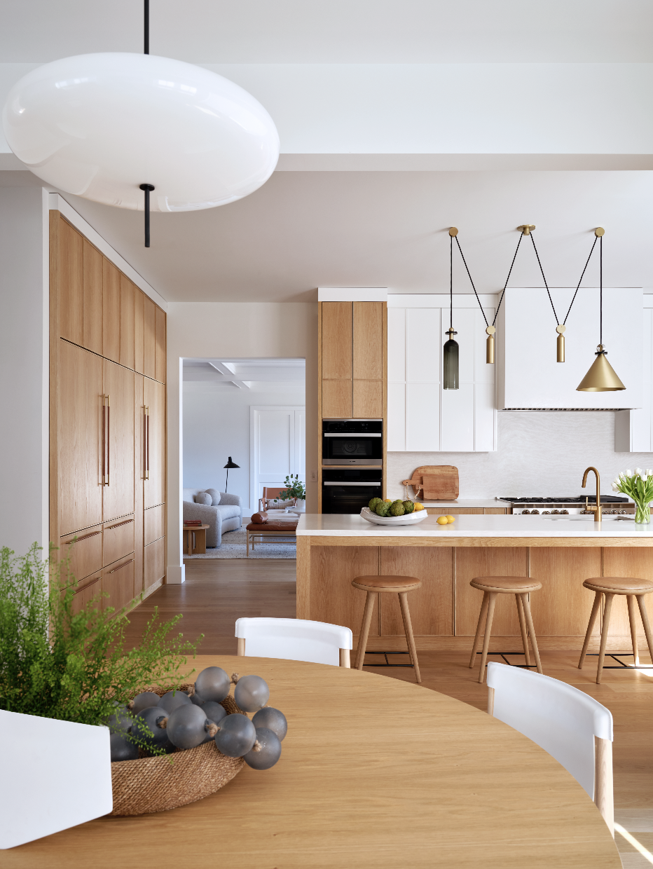 Hamptons Interior Design. Modern kitchen with wooden cabinets, white countertops, and gold fixtures, adjacent to a dining table with a plant and decorative objects.