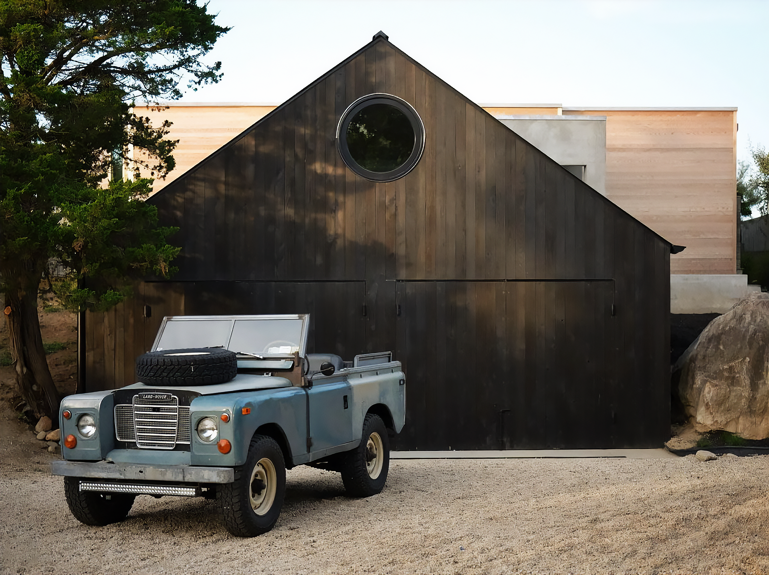 Hamptons Interior Design. A vintage Land Rover parked in front of a modern dark wooden and light beige house with a circular window near the peak of the roof, surrounded by gravel ground, large rocks, trees, and a partly cloudy sky.