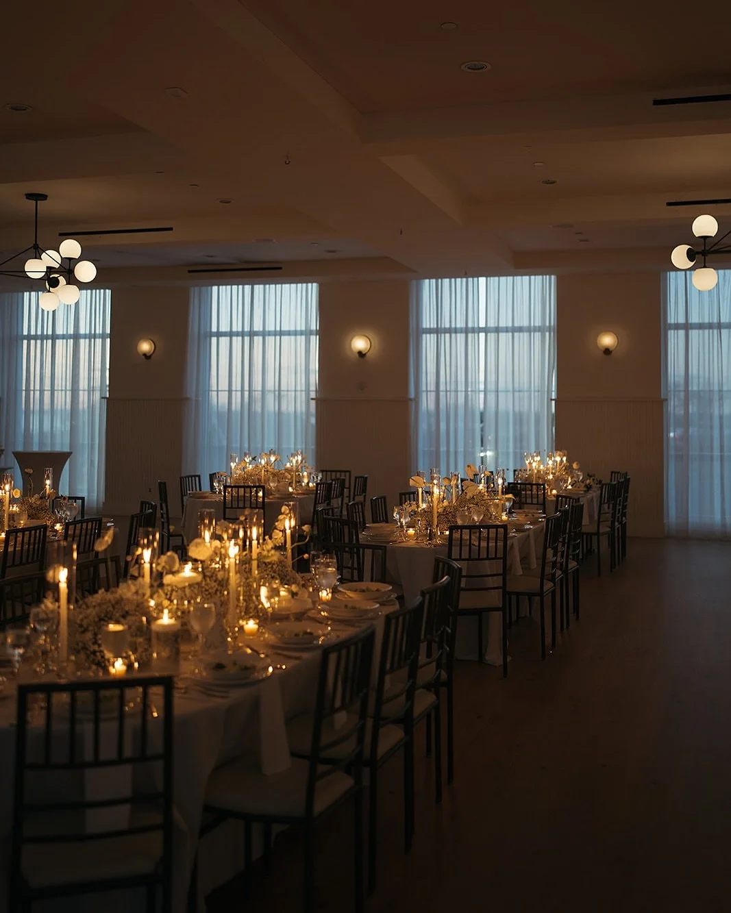Elegant banquet hall with round tables decorated with white tablecloths, floral centerpieces, and candlelights, set for an event, illuminated by soft lighting from wall sconces and hanging chandeliers, with large windows draped with sheer curtains.
