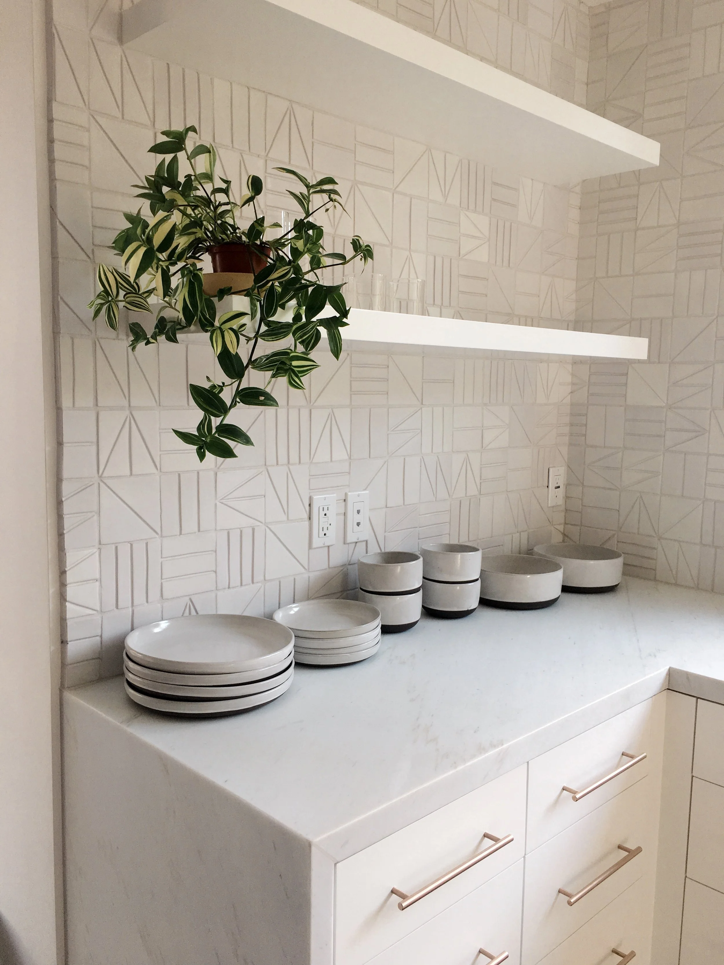 Los Angeles interior Design. Kitchen counter with white dishes, a potted plant, and two white shelves mounted on a geometric-patterned wall.