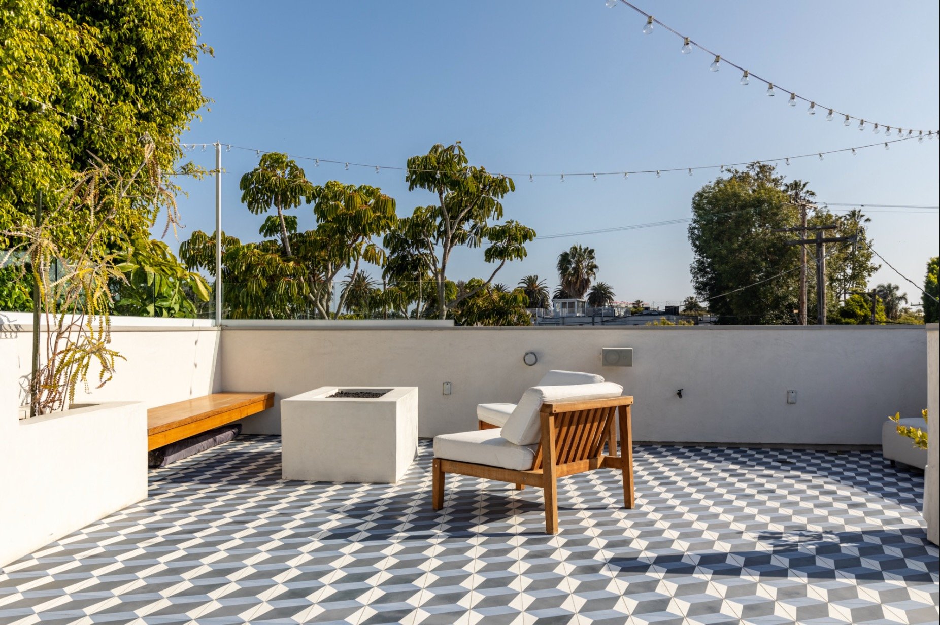 Los Angeles Roof Design. Outdoor patio with geometric black and white tile floor, white wall, two wooden chairs with white cushions, built-in bench, string lights overhead, and green trees in the background under a clear blue sky.