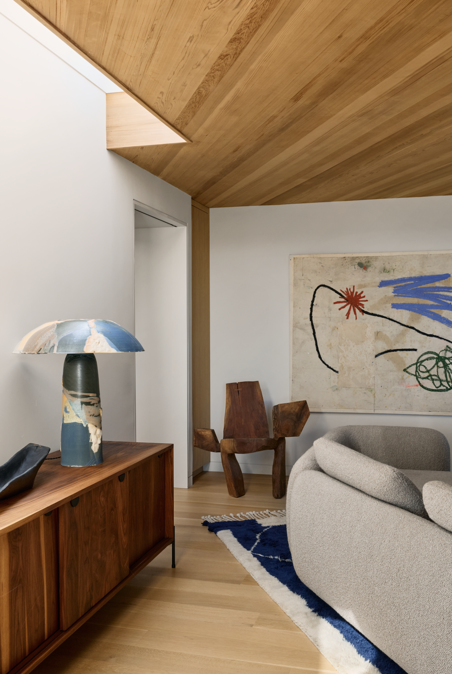 Hamptons Interior Design. Interior of a modern living room with a wooden ceiling, abstract artwork, a beige sofa, a wooden chair, a blue and white rug, and a decorative table lamp on a wooden sideboard.