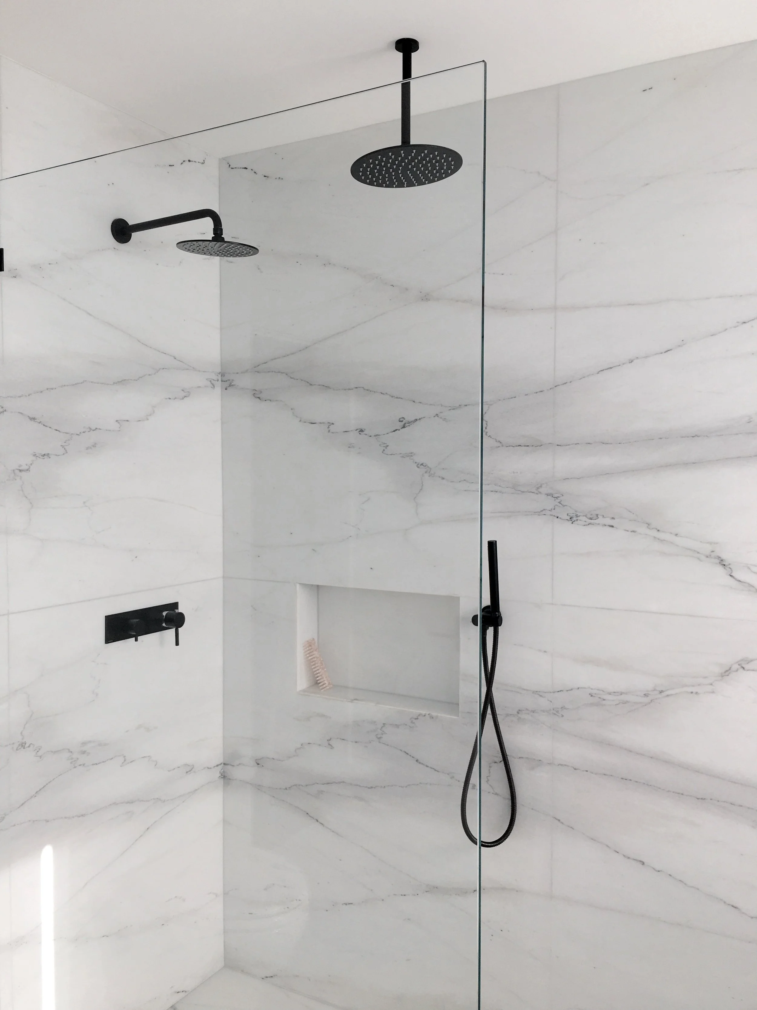 Los Angeles interior Design. Primary bathroom design. Modern walk-in shower with white marble walls, black fixtures, overhead rainfall showerhead, and a handheld showerhead, featuring a built-in shelf with a pink bath brush.
