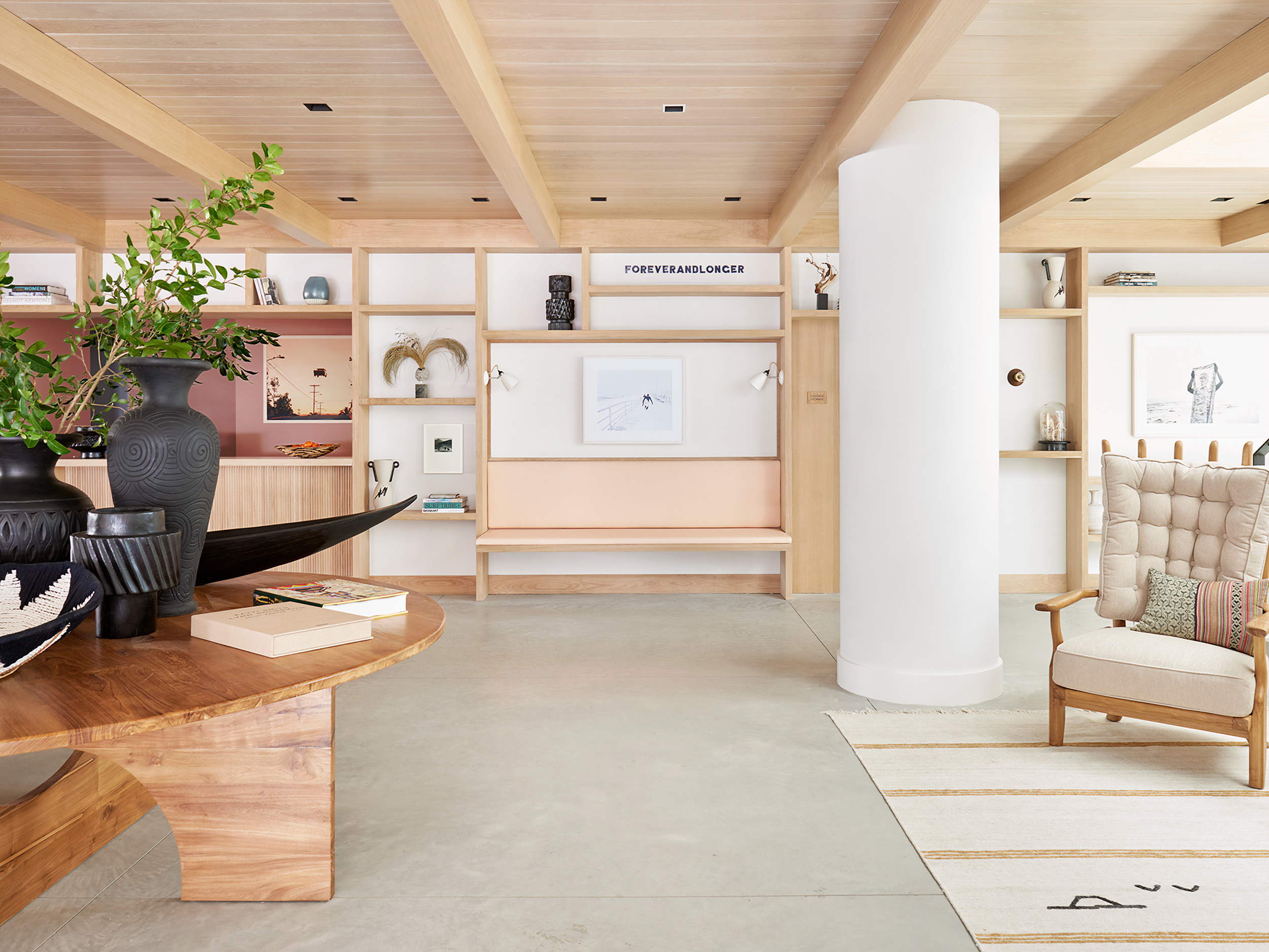 Modern living room with wooden ceiling beams, white walls, a round pillar, beige armchair, wooden shelving, and decorative items.