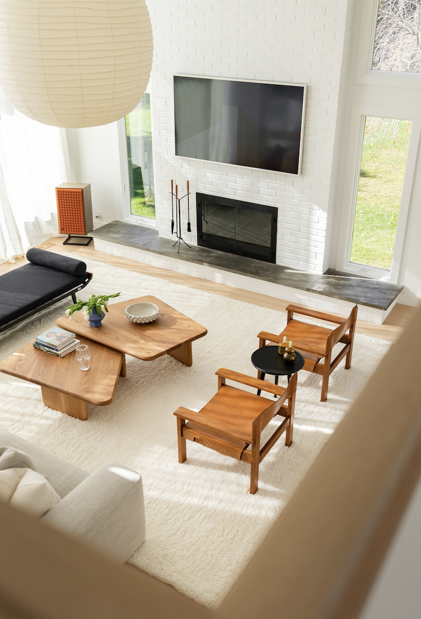 Hamptons Interior Design. Bright living room with white brick fireplace, wall-mounted TV, large windows with outdoor view, beige sofa, wooden coffee table, two wooden chairs with black side table, potted plant, books, glass, and decorative items.