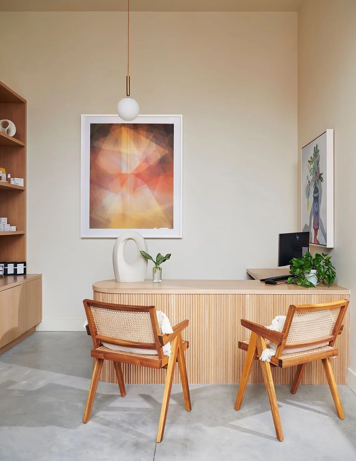 Minimalist office or reception area with a wooden desk, two wooden chairs with woven seats, a black computer monitor, green plants, and abstract artwork on the walls.