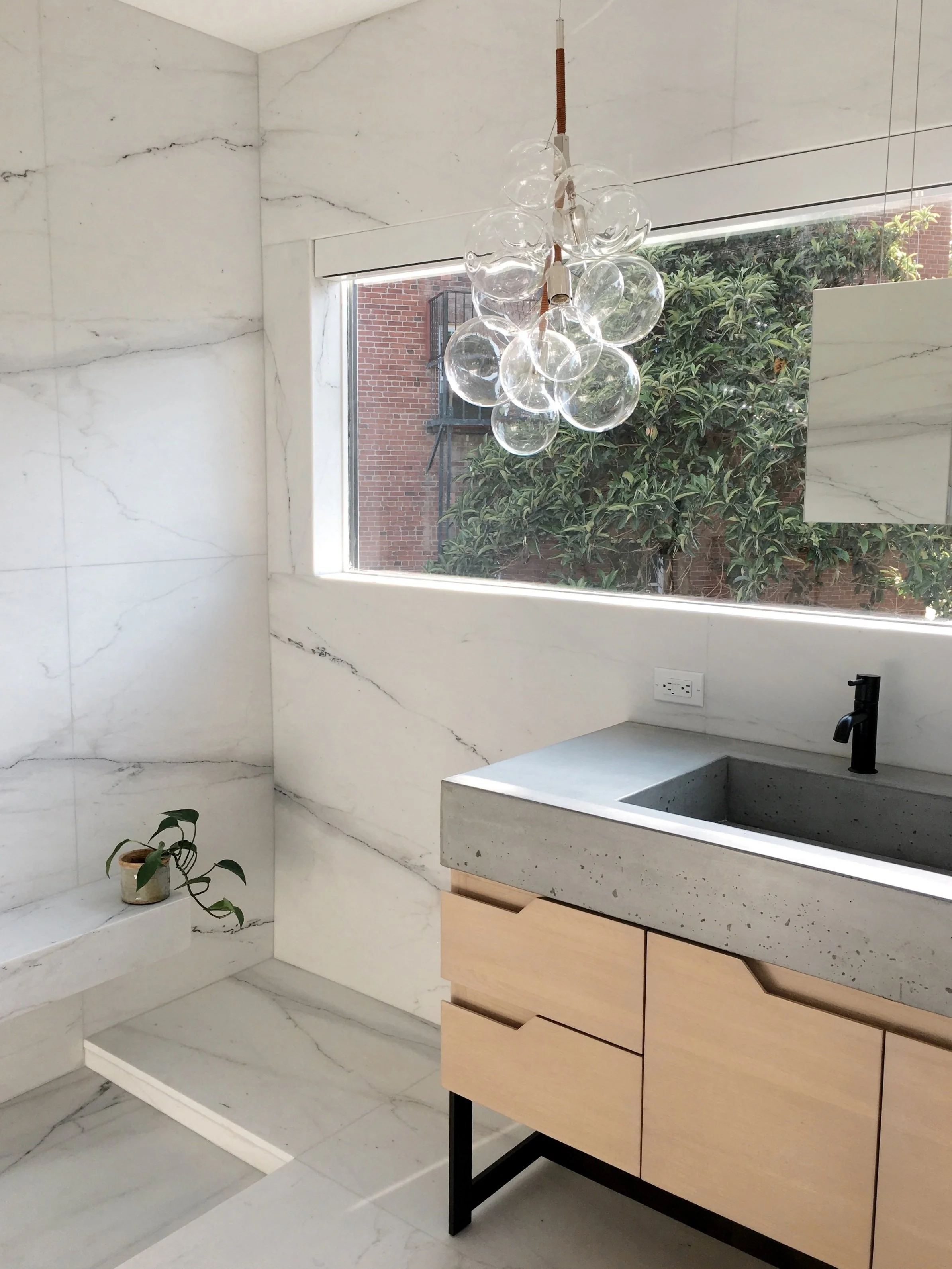 Los Angeles interior Design. Modern bathroom with marble walls and floors, a large window with greenery outside, a concrete sink with a black faucet, a wooden cabinet, a pendant light with glass globes, and a small potted plant on a marble shelf.