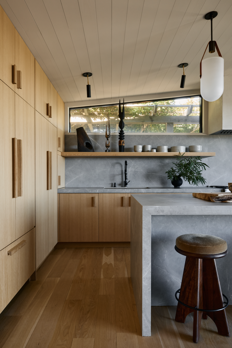 Modern kitchen with wooden cabinets, light gray stone countertops, a window above the sink, and decorative items including sculptures and bowls on a wooden shelf. There is a marble countertop island with a wooden stool and a potted plant nearby.