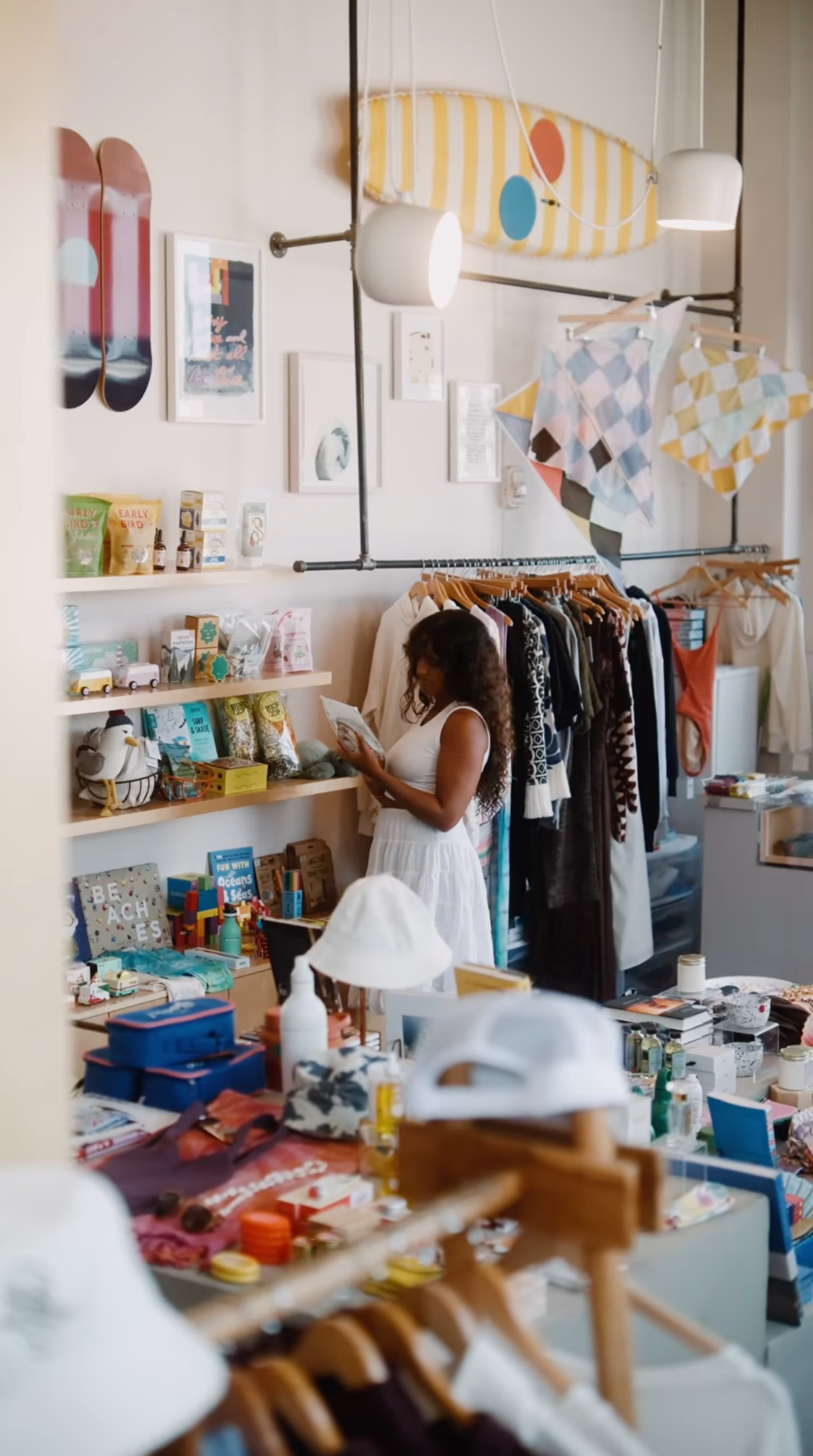 A woman with long curly hair wearing a white dress browsing items in a boutique store filled with clothing, accessories, and decorative items.