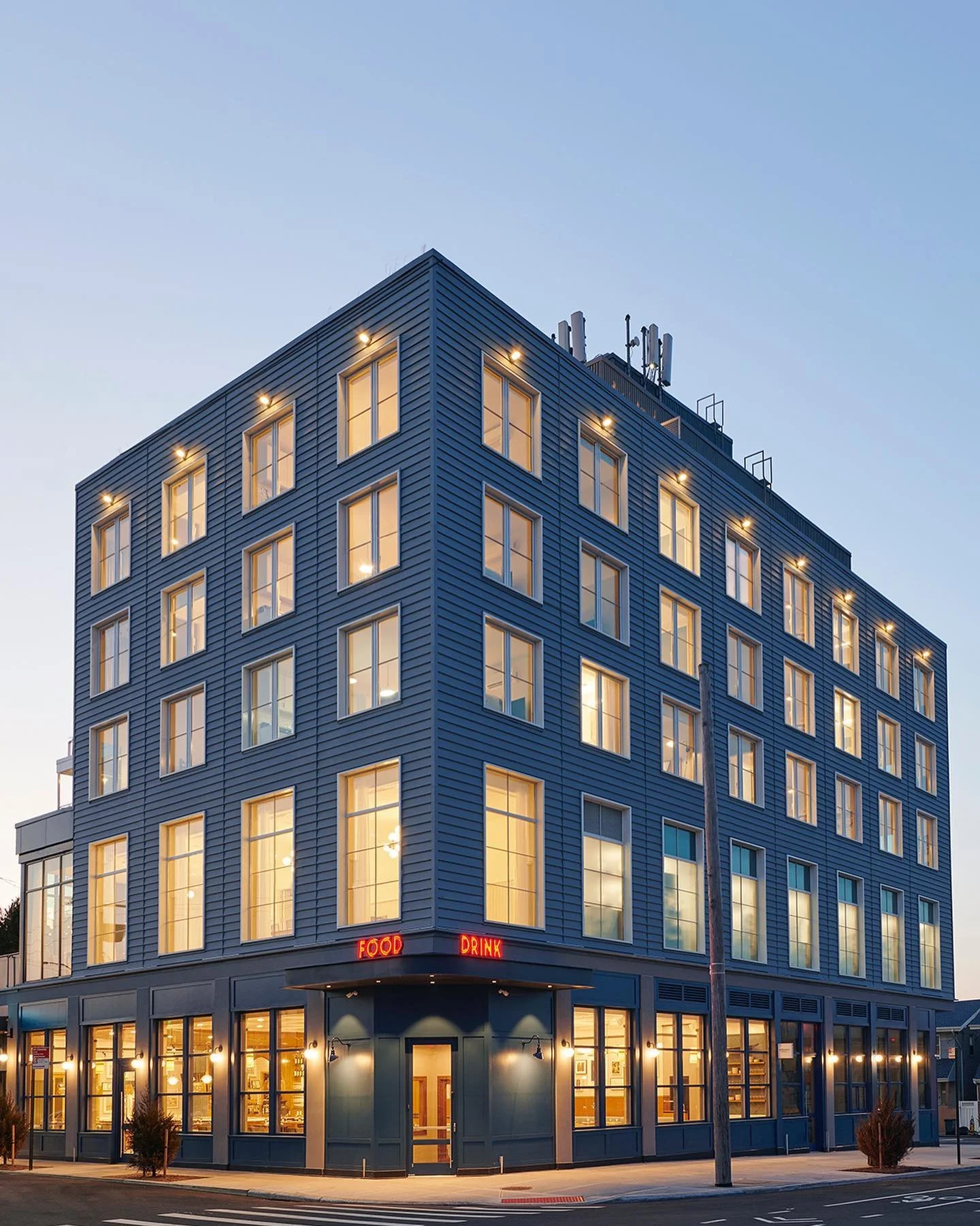 A multi-story modern building with a blue exterior, illuminated windows, and a neon sign that reads 'FOOD' and 'DRINK' at the corner.