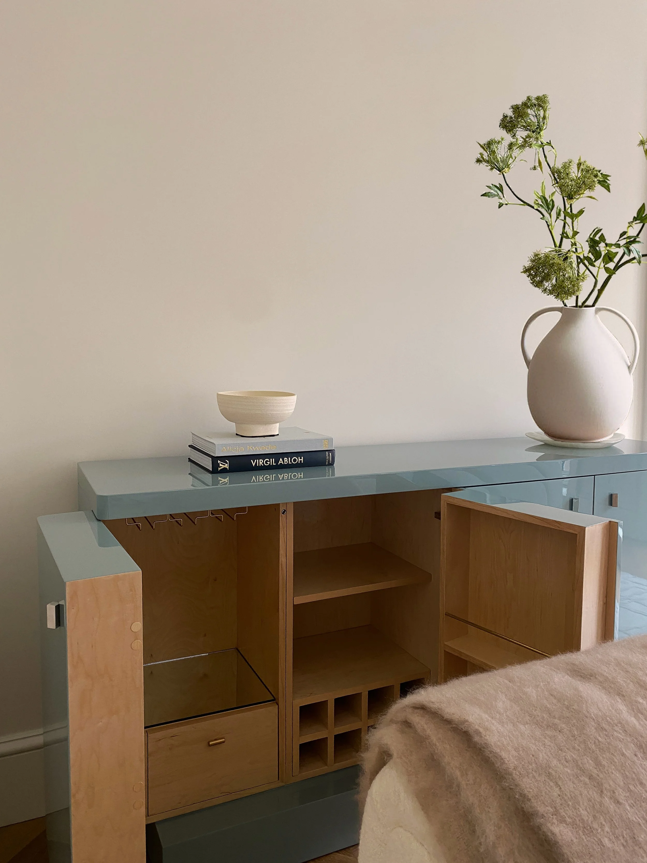 A modern sideboard with a pale blue top and wooden compartments, decorated with a tall white vase of green foliage, a white bowl, and a stack of two books.