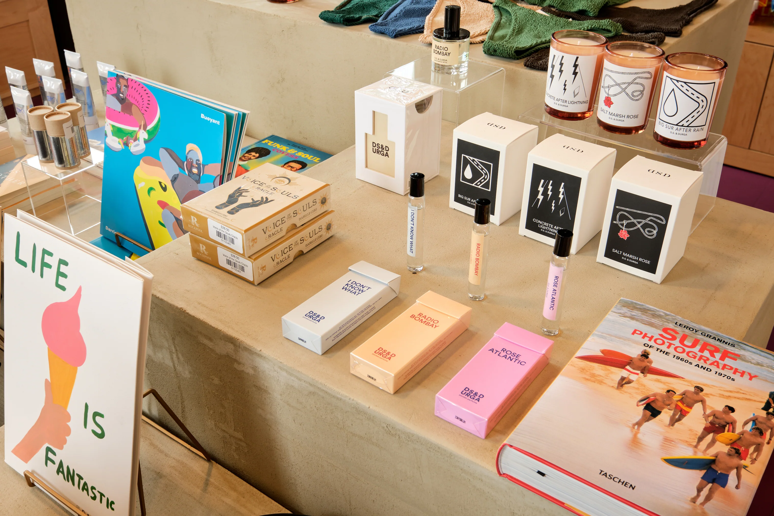 Display of various colorful greeting cards, scented candles, small perfume bottles, and books on a beige table at a store. The greeting cards have playful designs including an ice cream cone with the text 'Life is Fantastic' and another with illustrated animals and the word 'Buoyant.' The candles are in small glass jars with designs and names such as 'Salt Marsh Rose' and 'Big Sur After Rain.' There are perfume vials labeled 'Radio Bombay,' 'Rose Atlantic,' 'I Don't Know What,' and 'Concrete After Lightning.' A large book titled 'Surf Photography of the 1960s and 1970s' lies on the table, and behind it are folded clothes or towels.