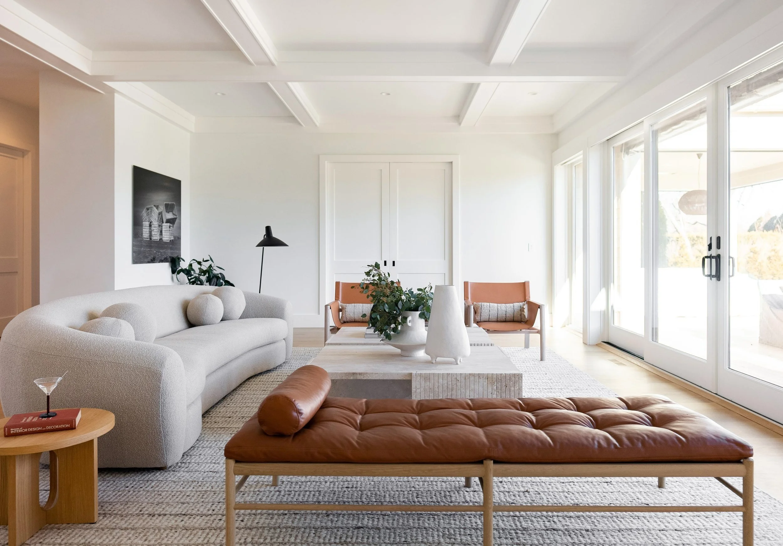 Bright, modern living room with large glass sliding doors, white walls, and minimalist decor. Features a white sofa with rounded pillows, a leather bench, a wooden side table with a martini glass, and a black floor lamp. A coffee table with decorativ