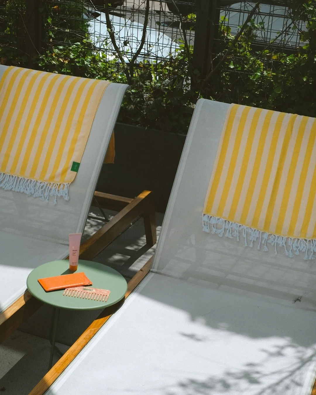 Two white lounge chairs with yellow and white striped towels draped over them, a small round green table with skincare products and an orange wallet, and a fence with greenery in the background, suggesting an outdoor pool area.