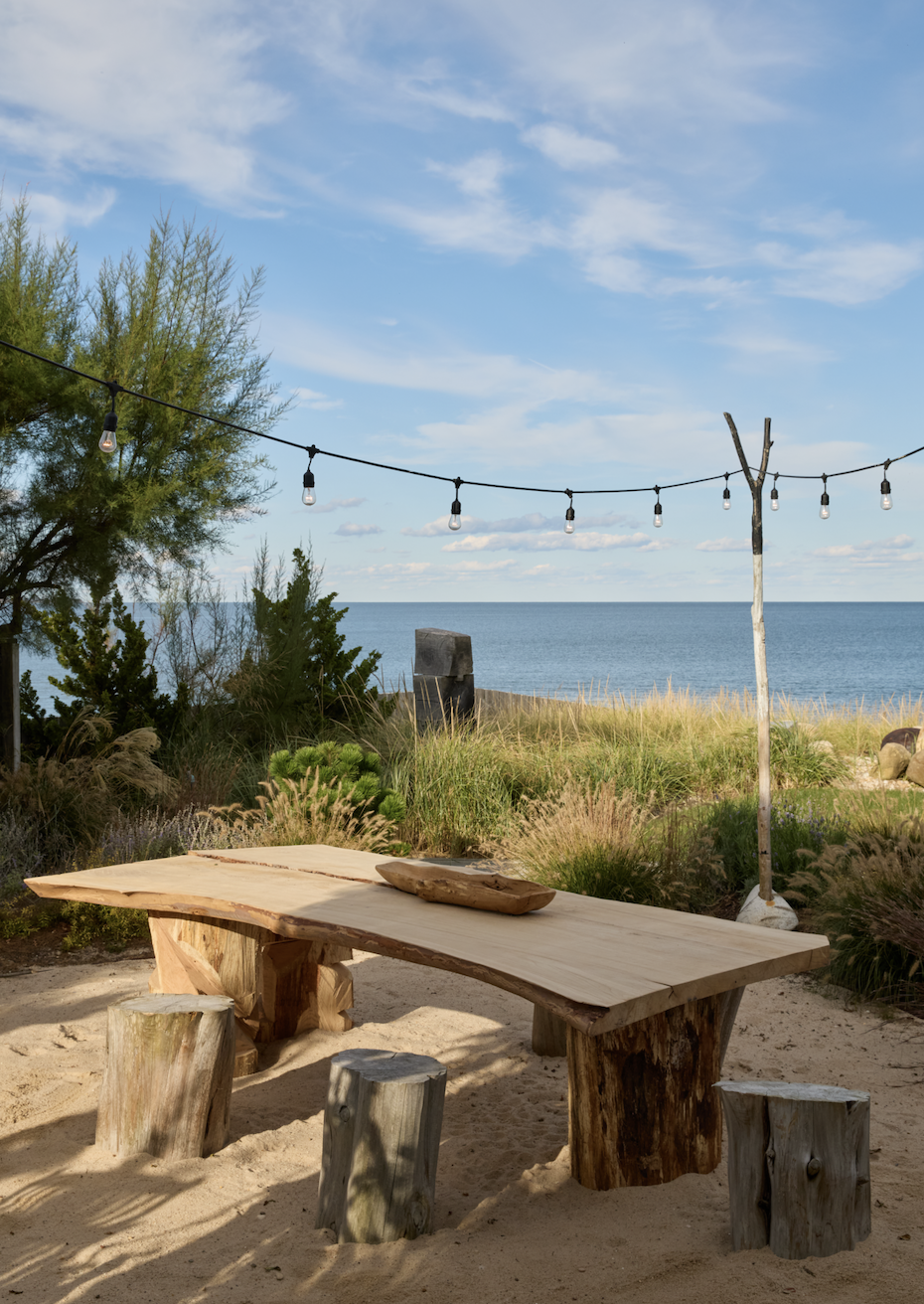 A rustic wooden outdoor table with matching stools, situated on sandy ground near a beach, with string lights overhead, greenery around, and the ocean in the background under a blue sky with scattered clouds.