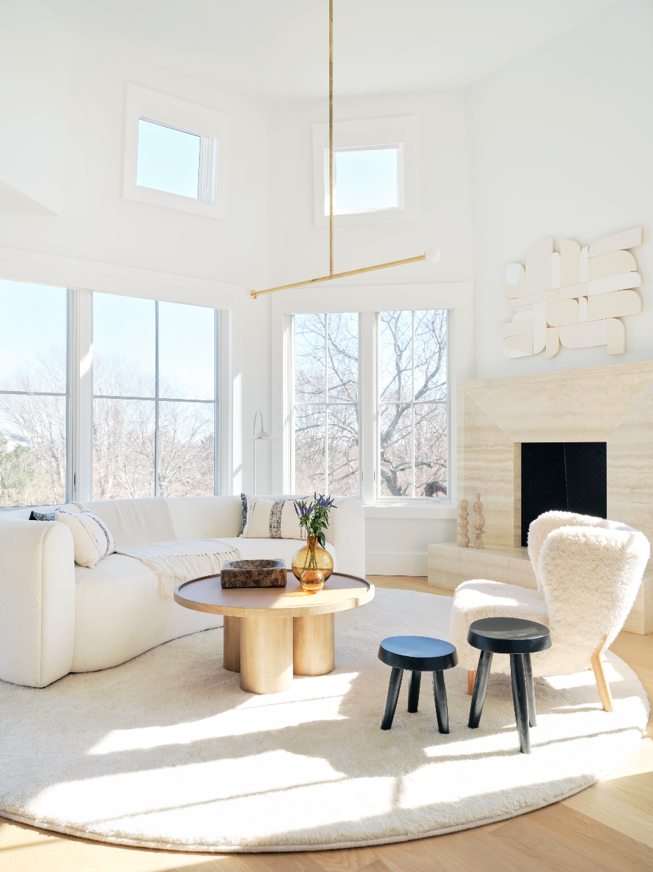 New York Interior Design. Hamptons Design. Bright bedroom sitting area with large windows, white sofa, round wooden coffee table, plush white chair, black stools, and contemporary art above fireplace.