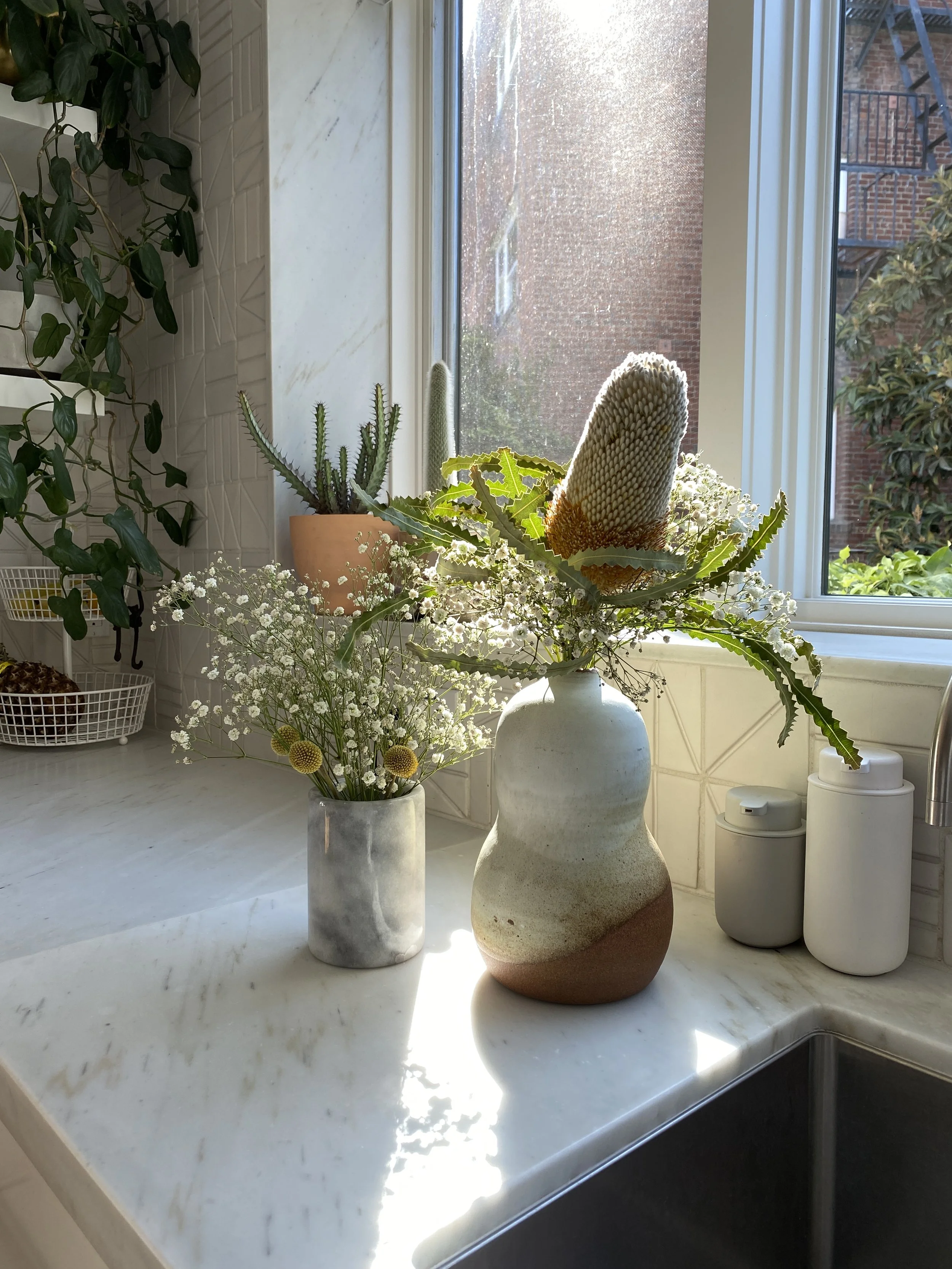 Los Angeles interior Design. Vase with white, yellow, and green flowers, and a smaller marble vase with white daisies, on a kitchen countertop by a window with sunlight.