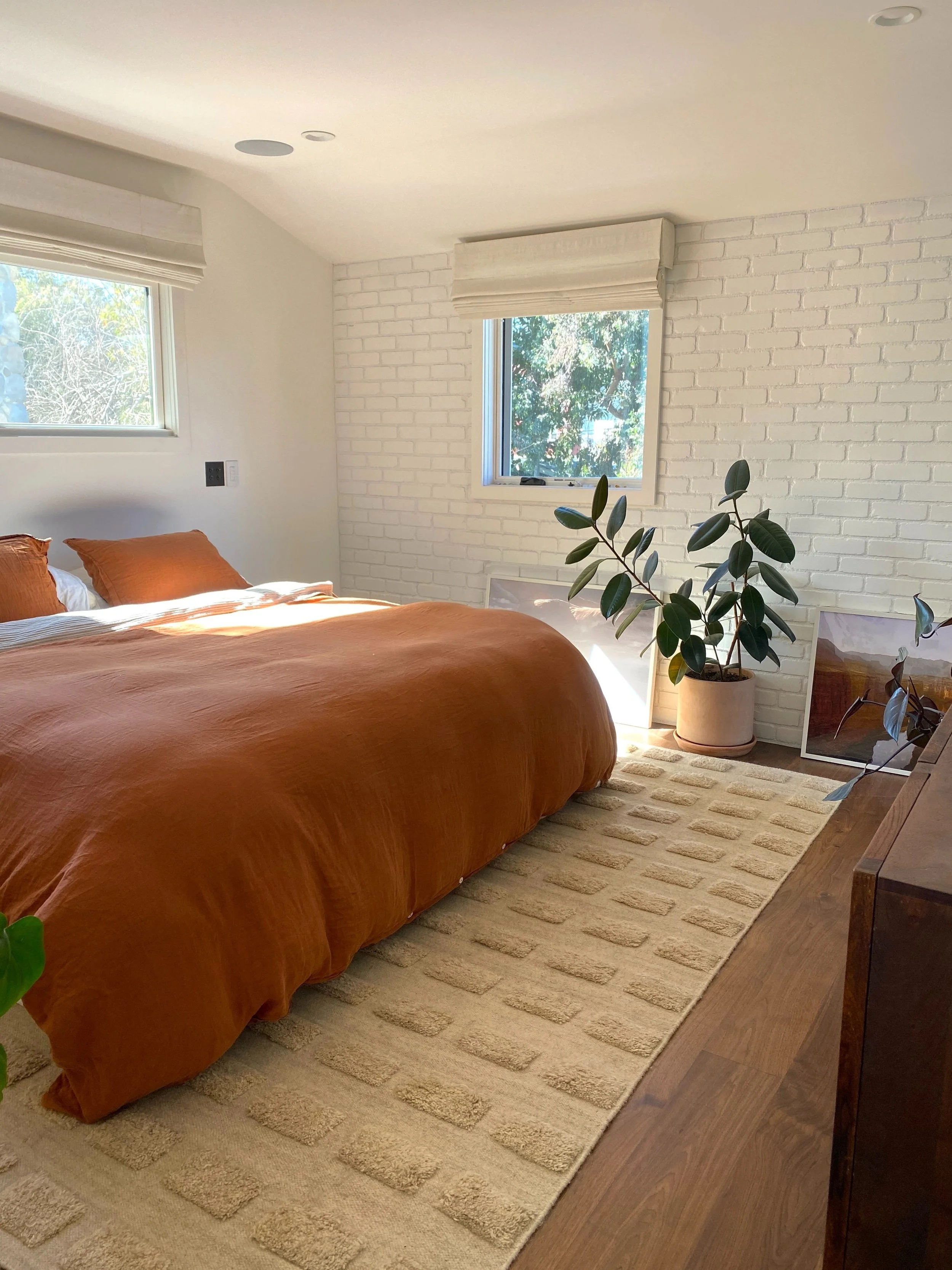 Los Angeles interior Design. A bedroom with a bed covered in an orange duvet, white walls, a white brick accent wall, two windows with shades, a large potted plant, framed pictures leaning against the wall, and a beige rug on a wooden floor.