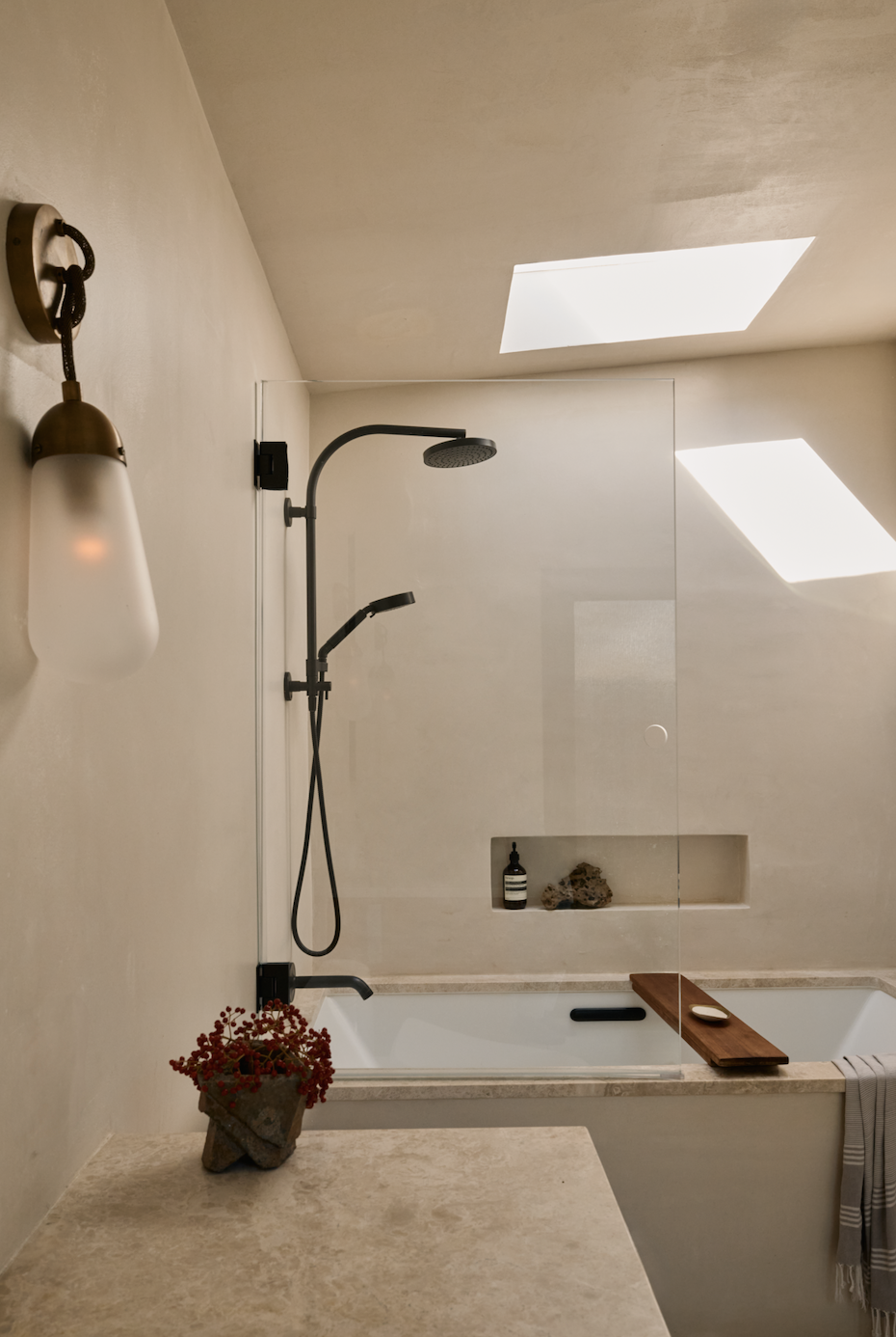 A minimalist bathroom with a bathtub, a black showerhead with a handheld attachment, a built-in shelf with toiletries, skylights letting in natural light, a wall-mounted light fixture, and a stone with a bottle of soap on the shelf. There's a plant on the counter and a towel hanging on the side of the bathtub.