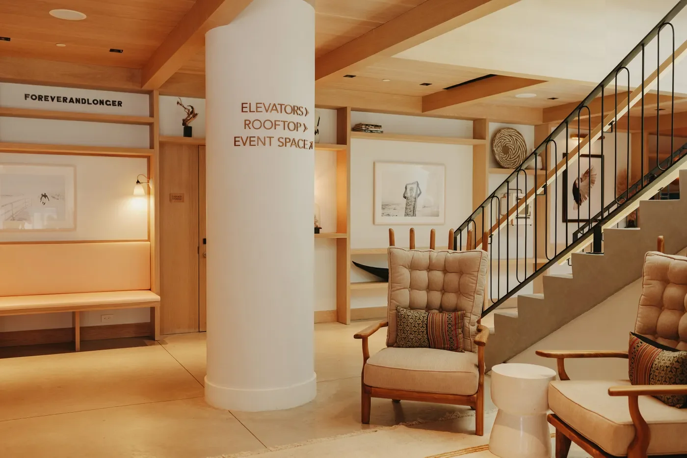 Interior of a lounge with wooden accents, armchairs, framed art on the walls, a staircase with black railing, and a white pillar with directional signs for elevators, rooftop, and event space.