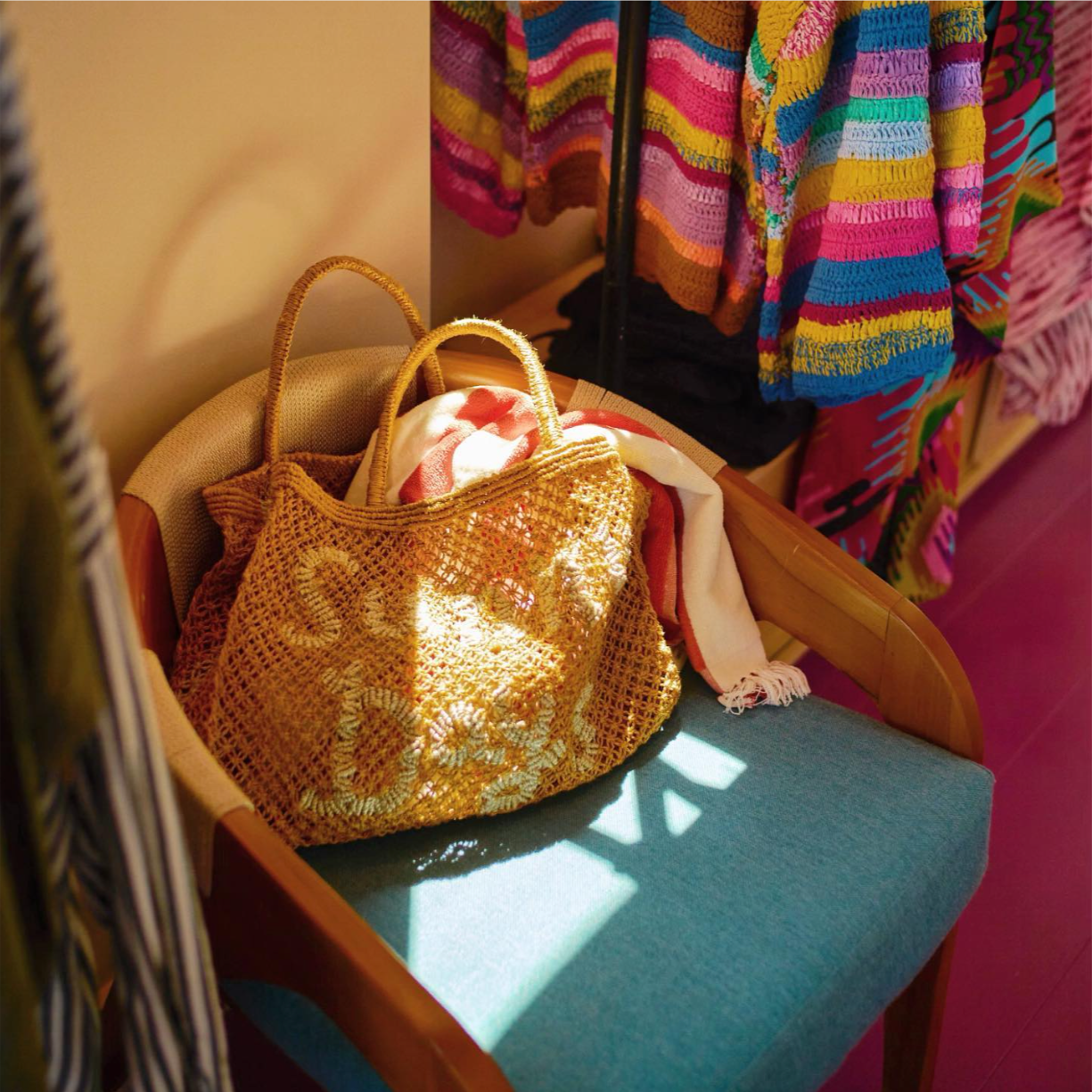 A woven straw tote bag with floral patterns resting on a teal cushioned chair, with a white and red striped towel spilling out. Behind, vibrant multicolored knitted garments and textiles are displayed.