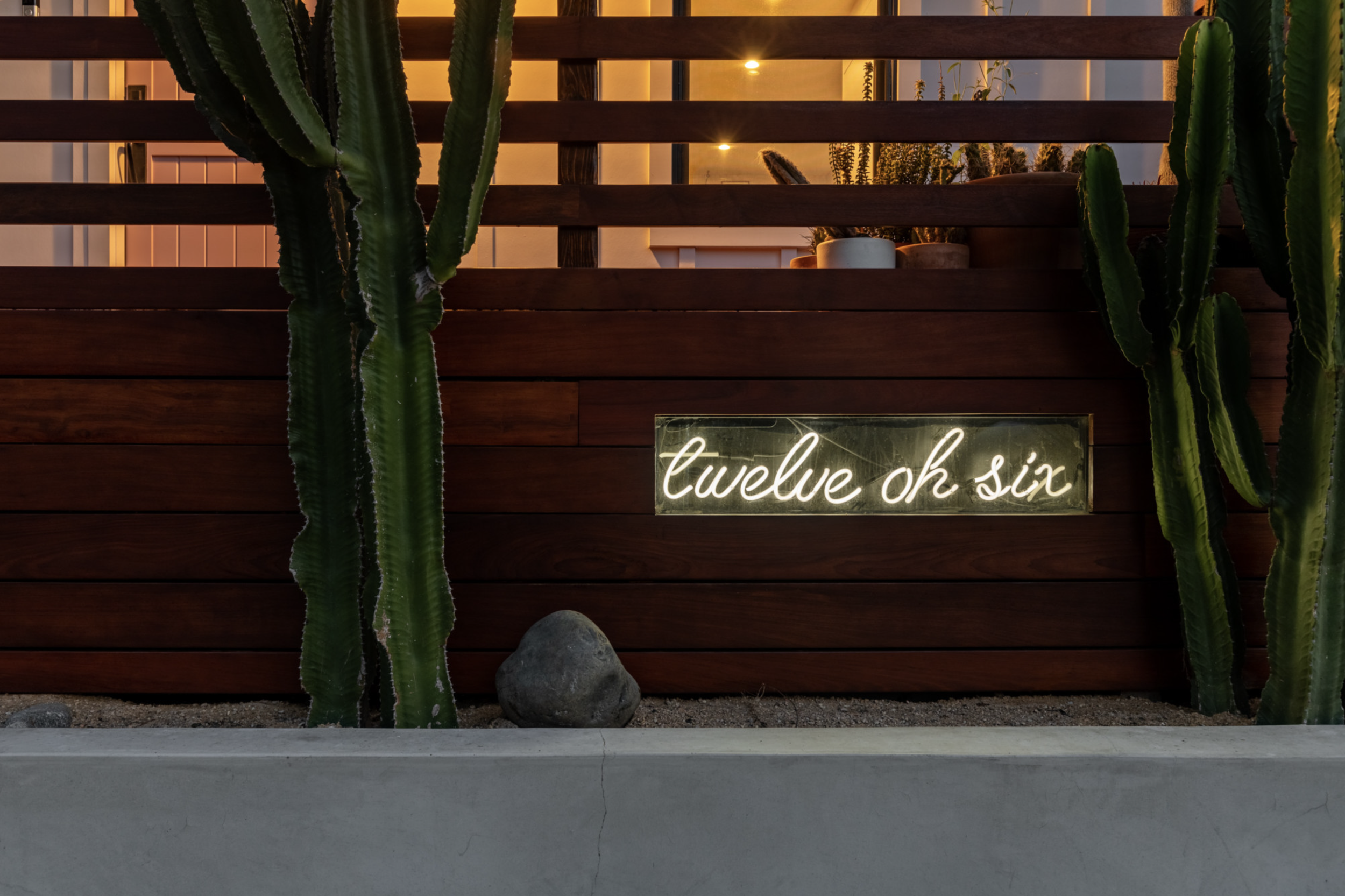 Neon house sign. A wooden fence with green cacti and a neon sign that reads "twelve oh six" in cursive. The scene is set at dusk with warm lighting.