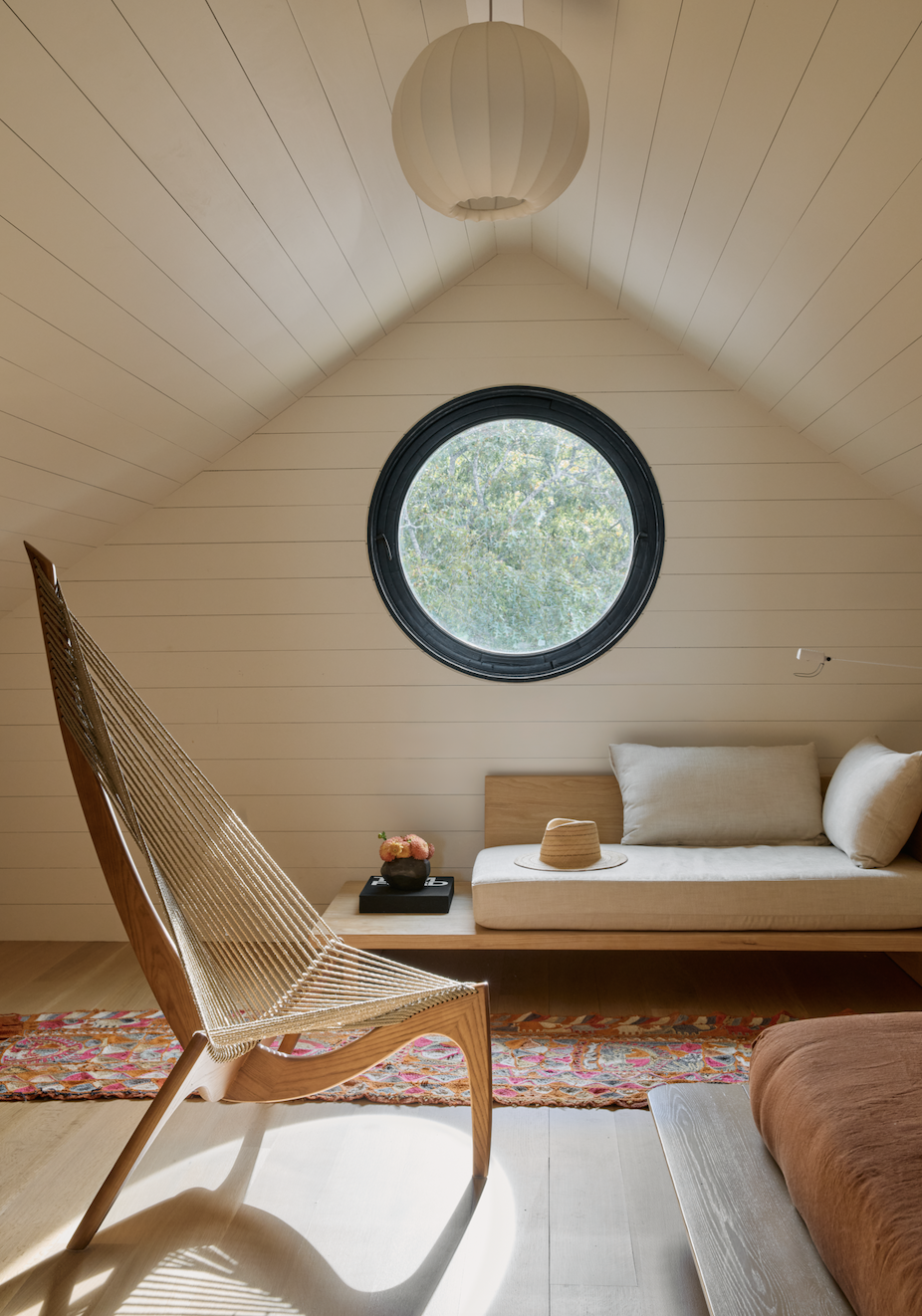 Hamptons Interior Design. Interior of a cozy room with a sloped ceiling, featuring a circular window showing trees outside, a wooden bench with cushions, a modern wooden chair, a rug, and a paper lantern hanging from the ceiling.
