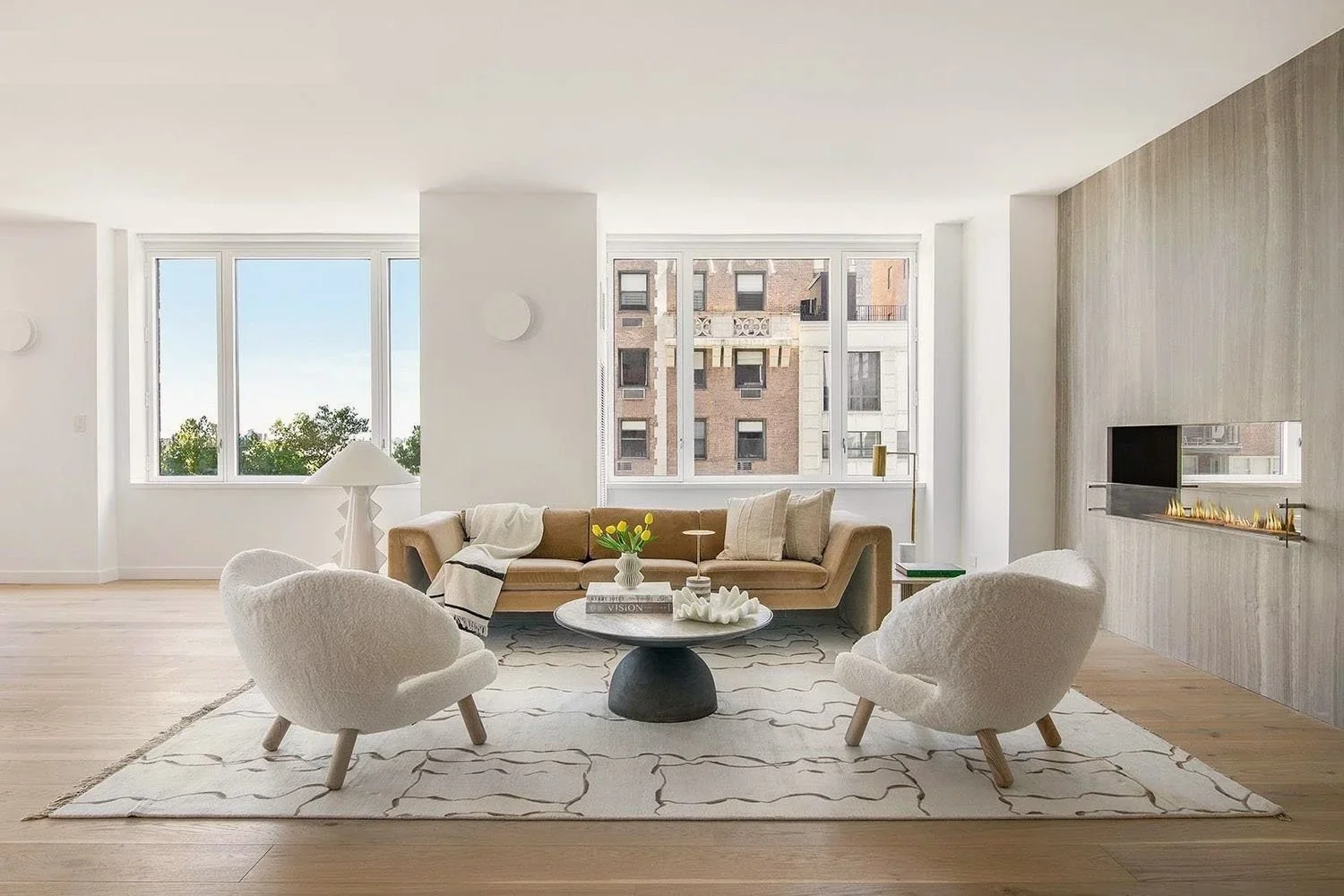 New York Interior Design. Modern living room with large windows, beige sofa, white armchairs, a round coffee table, and a fireplace, with a light wood floor and neutral decor.