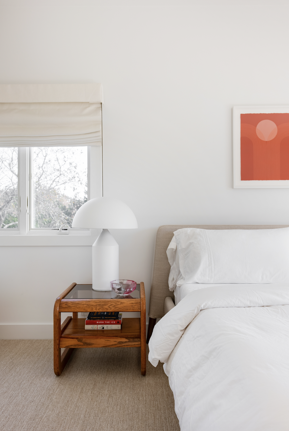 Hamptons Interior Design. A bedroom corner with a bed, a wooden nightstand, a white table lamp, a glass bowl, a window with a white blind, and a red abstract art piece on the wall.