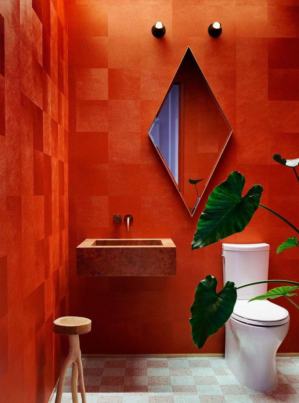 New York Interior Design. Powder room design. Modern bathroom with red textured walls, diamond-shaped mirror, indoor plants with large green leaves, a white toilet, a small wooden stool, a wall-mounted sink, and a window reflected in the mirror.