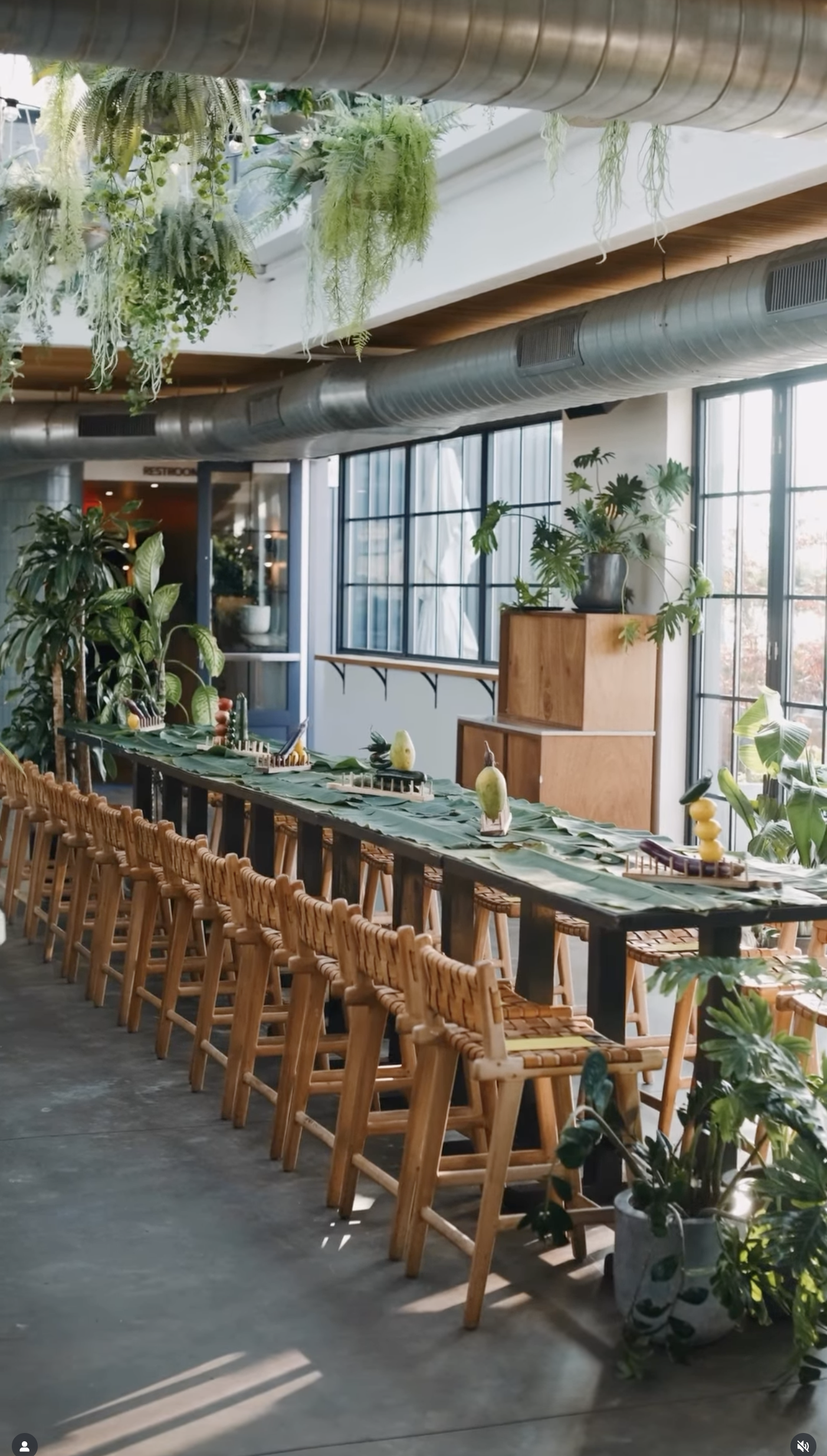 Rockaway Hotel Roof Restaurant. A long dining table decorated with green leaves and small fruit sculptures, surrounded by wooden chairs, inside a bright, modern space with lots of plants and large windows.