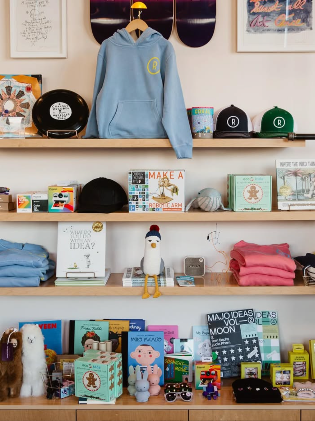 Display of various merchandise on wooden shelves, including a blue hoodie with a yellow 'R' logo, two baseball caps with 'R', plush toys, books, framed artwork, and decorative items.